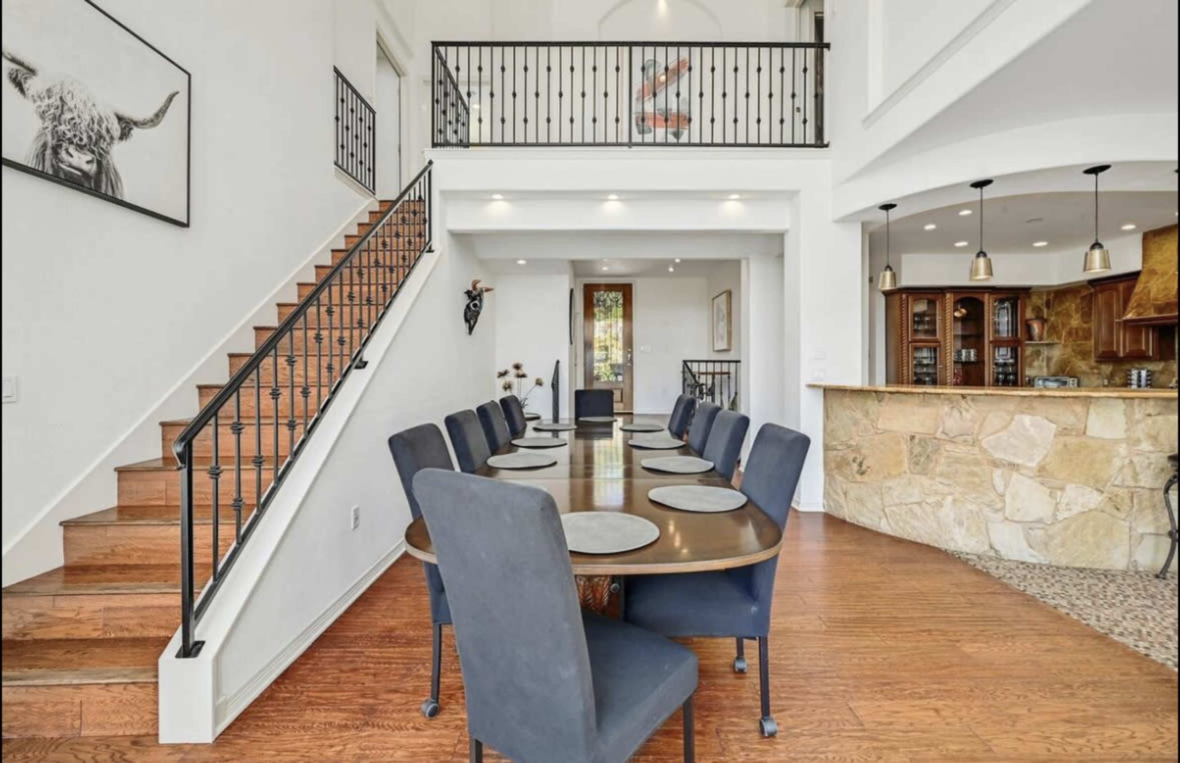A spacious dining area features a long table set with dark chairs, leading to a staircase and an open kitchen with a stone counter.