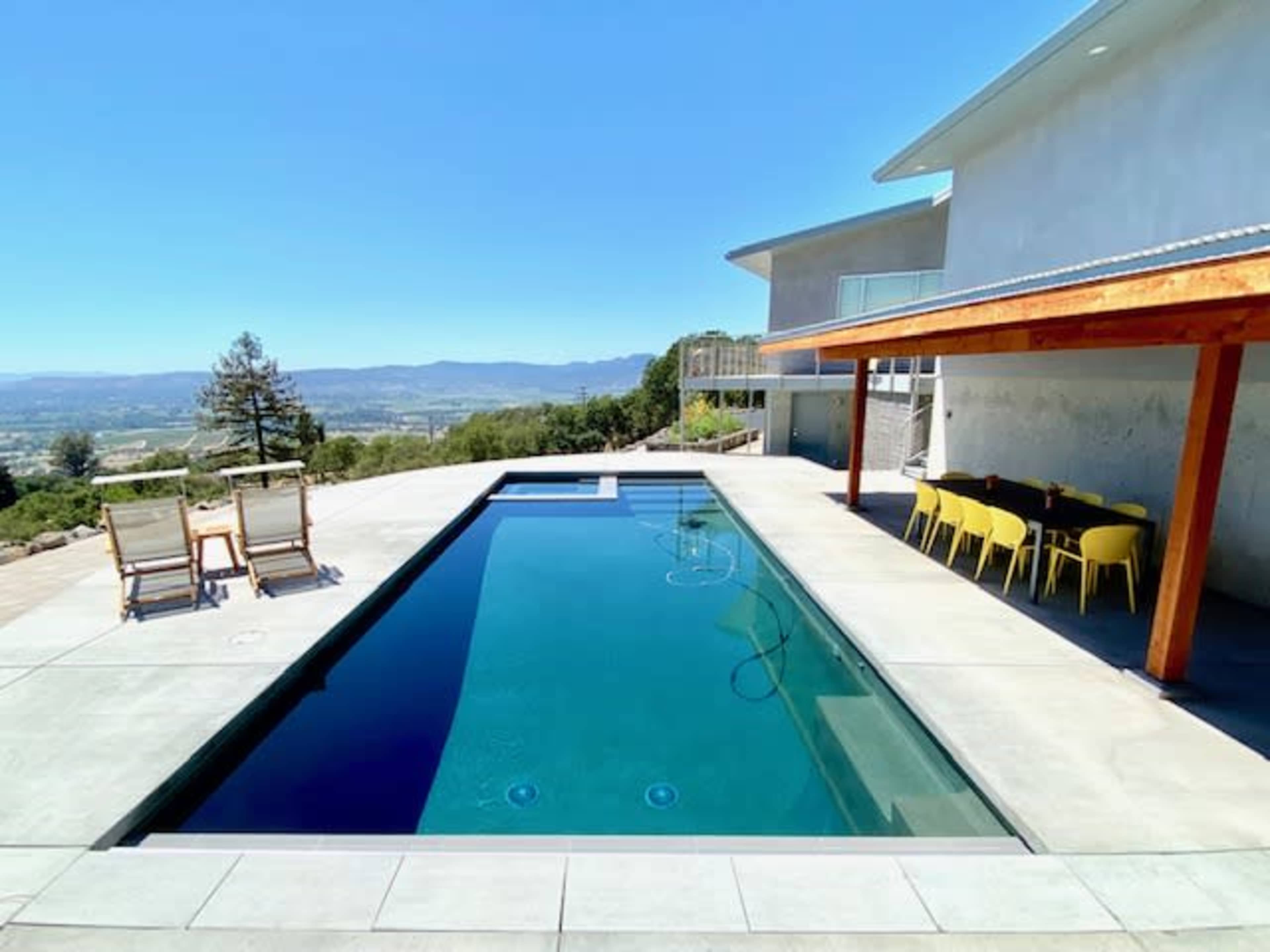 A modern backyard with an infinity pool, loungers, and a dining area under a wooden pergola, surrounded by a scenic landscape.