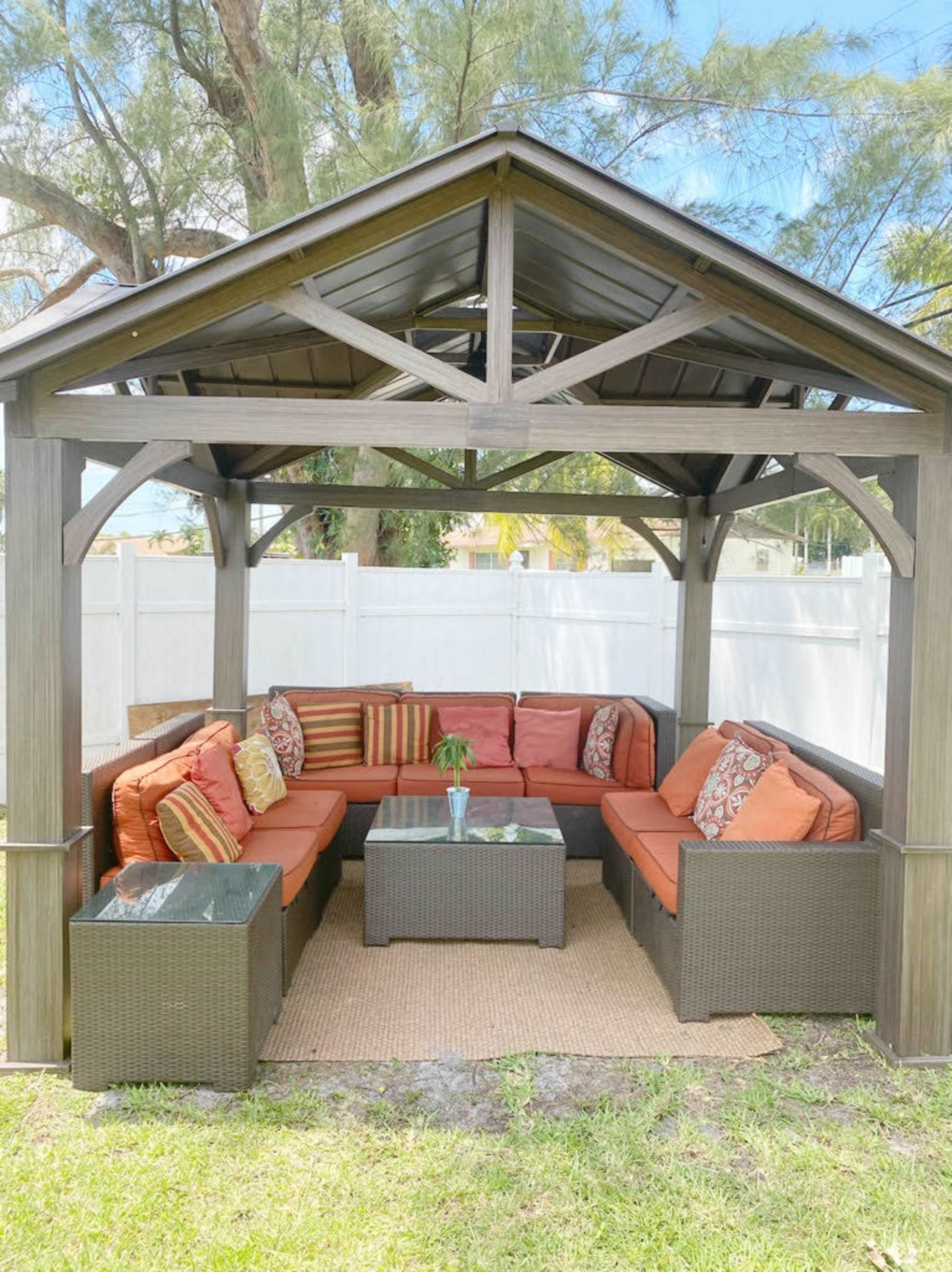 A covered outdoor seating area furnished with a sectional sofa, decorative cushions, and a coffee table, situated on a lawn next to a white fence.