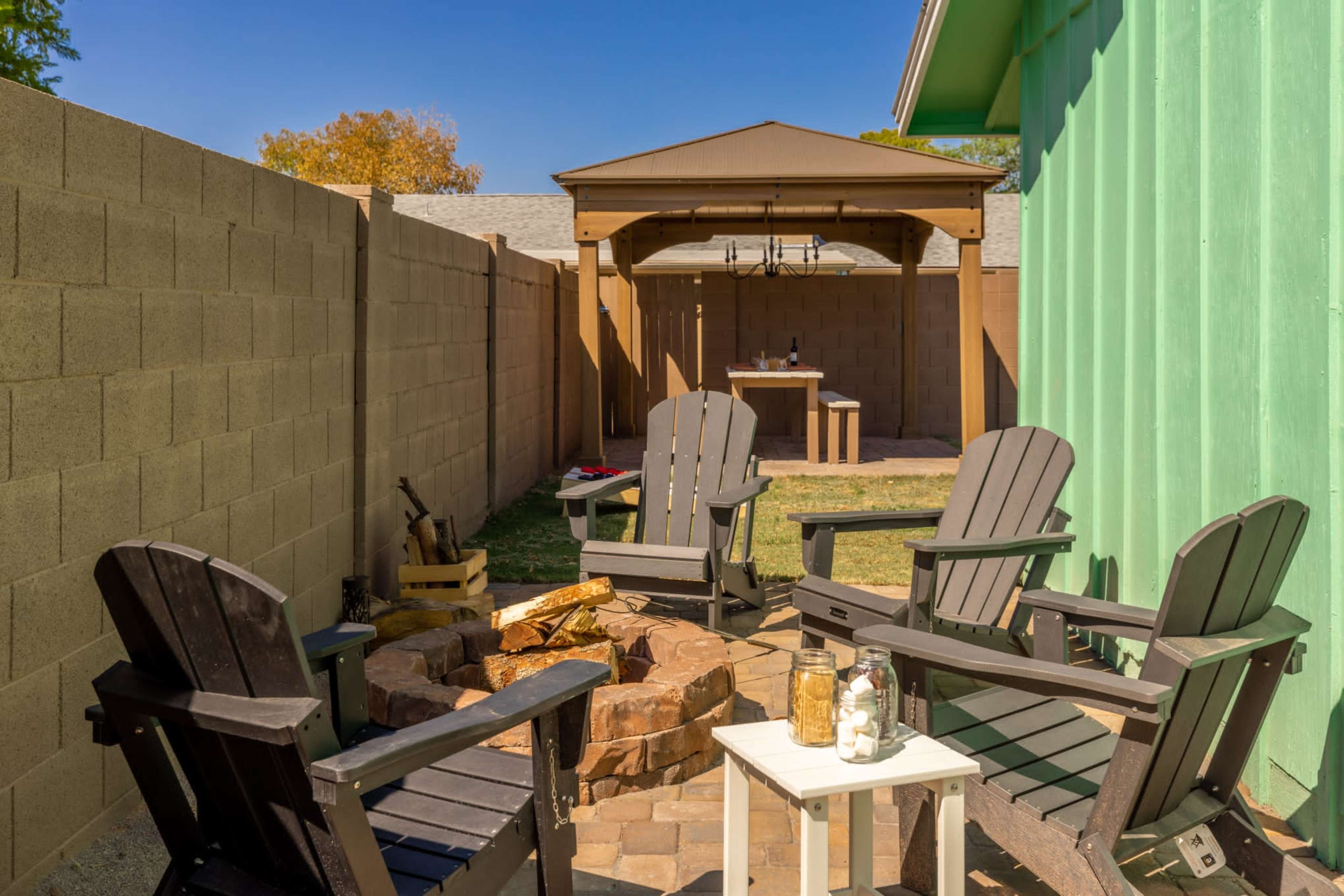 A backyard scene featuring a fire pit surrounded by four wooden chairs, with a gazebo and a table in the background.