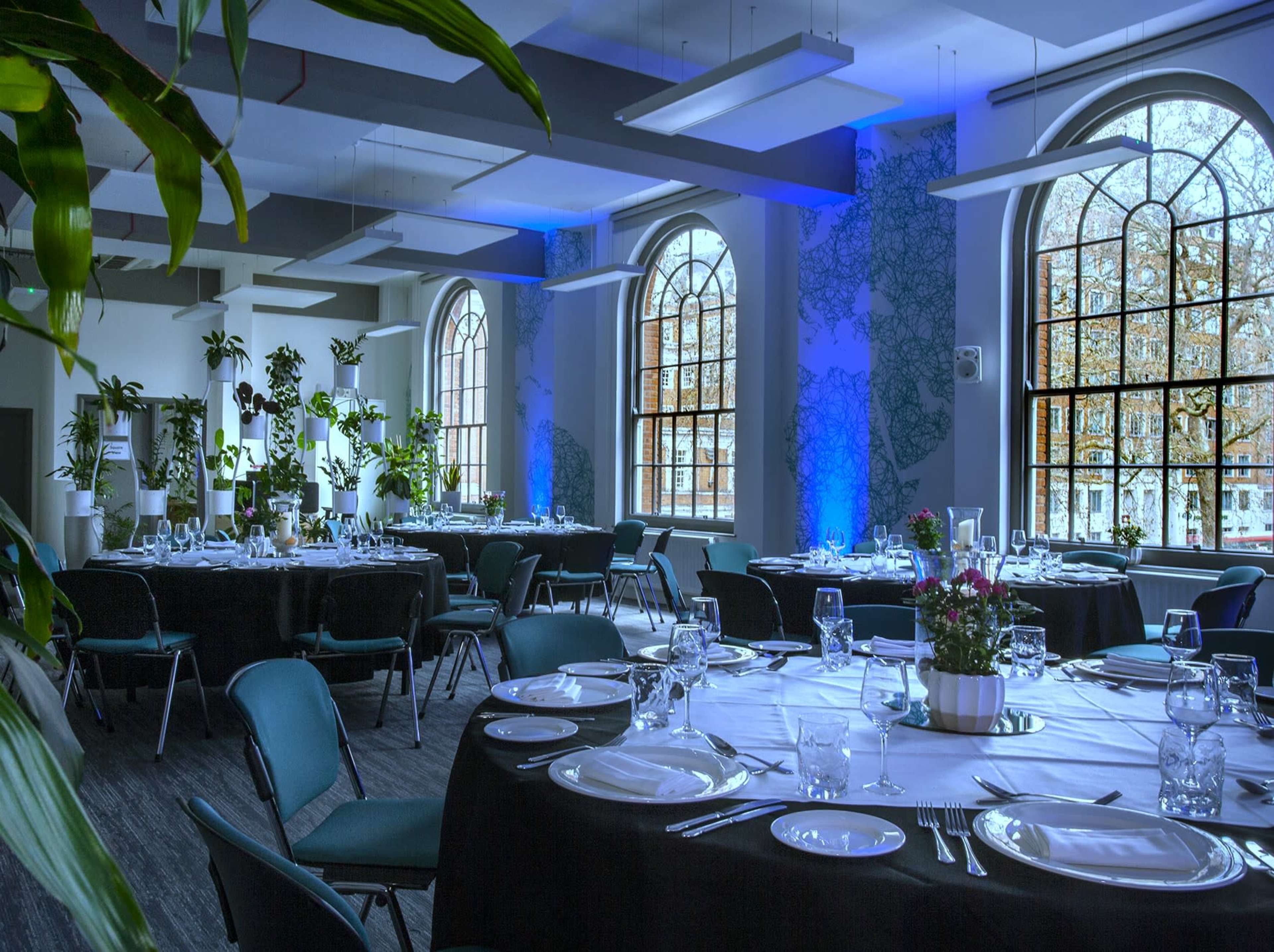 A modern dining room set for an event, featuring elegantly arranged tables with plates and glasses, surrounded by large windows and green plants.