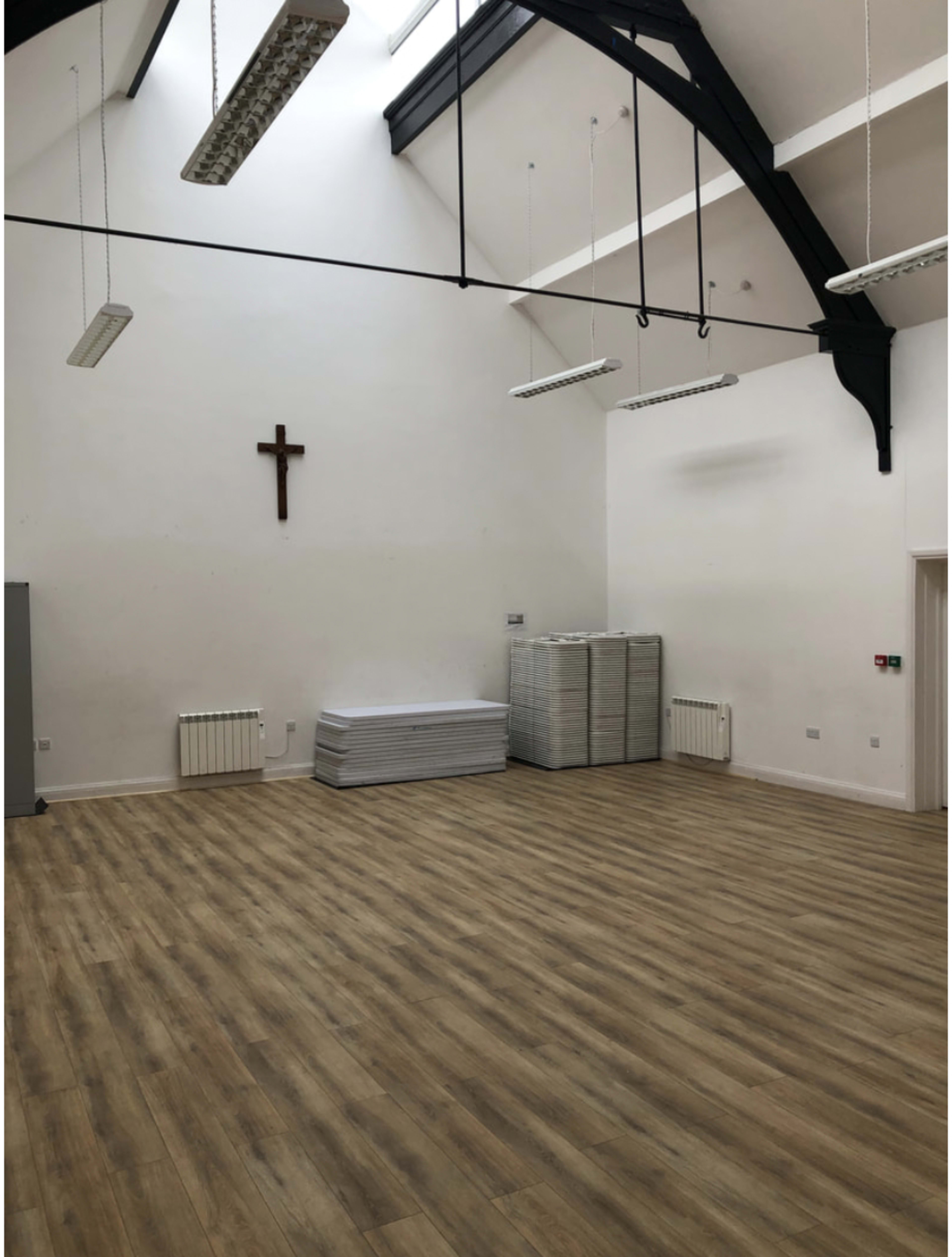 An empty hall with a wooden floor, featuring a cross mounted on the wall and stacks of folding tables in the corner.