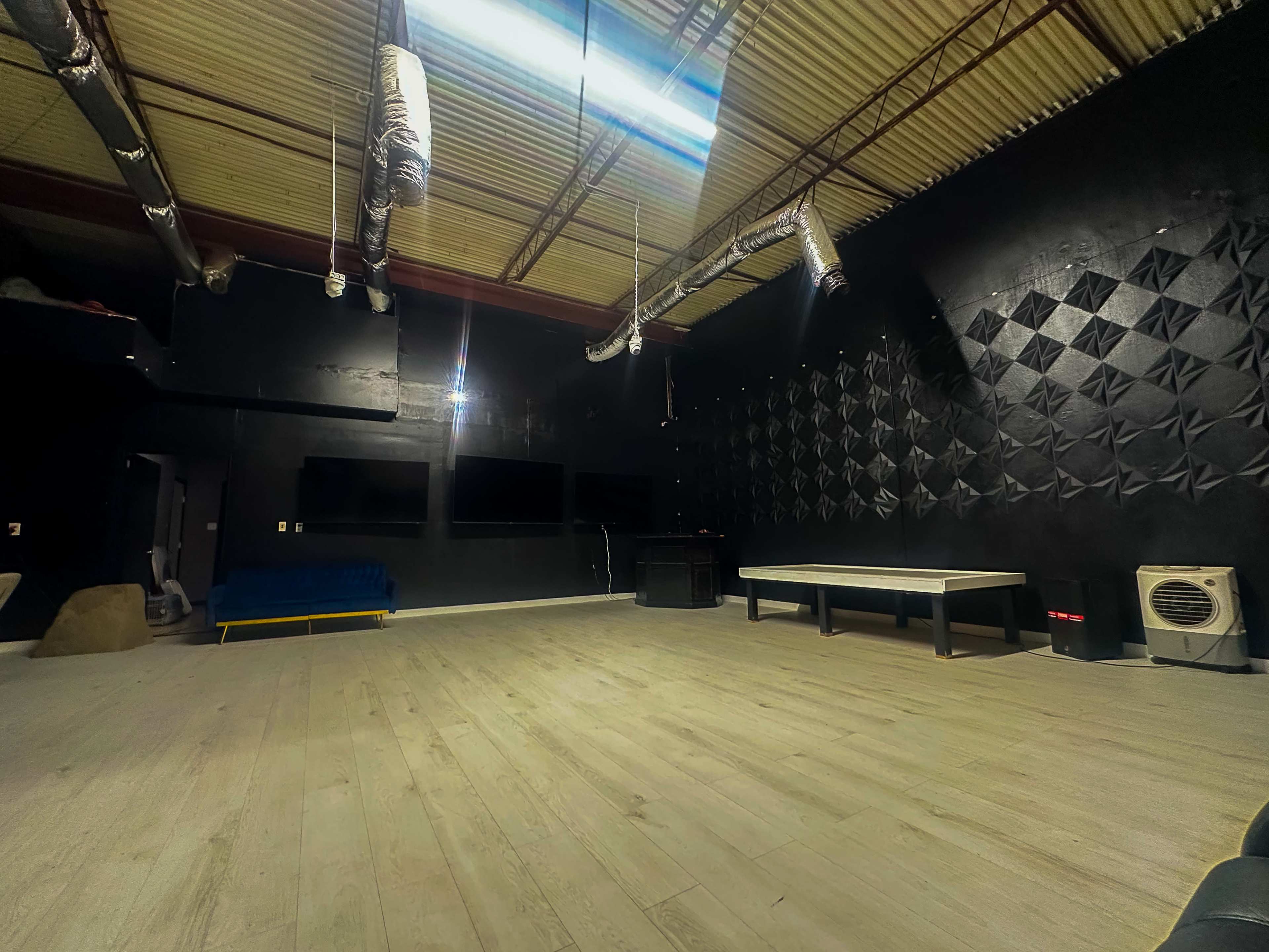 The image shows a large, empty room with black walls, a wooden floor, and minimal furnishings, including a white table and a blue couch.