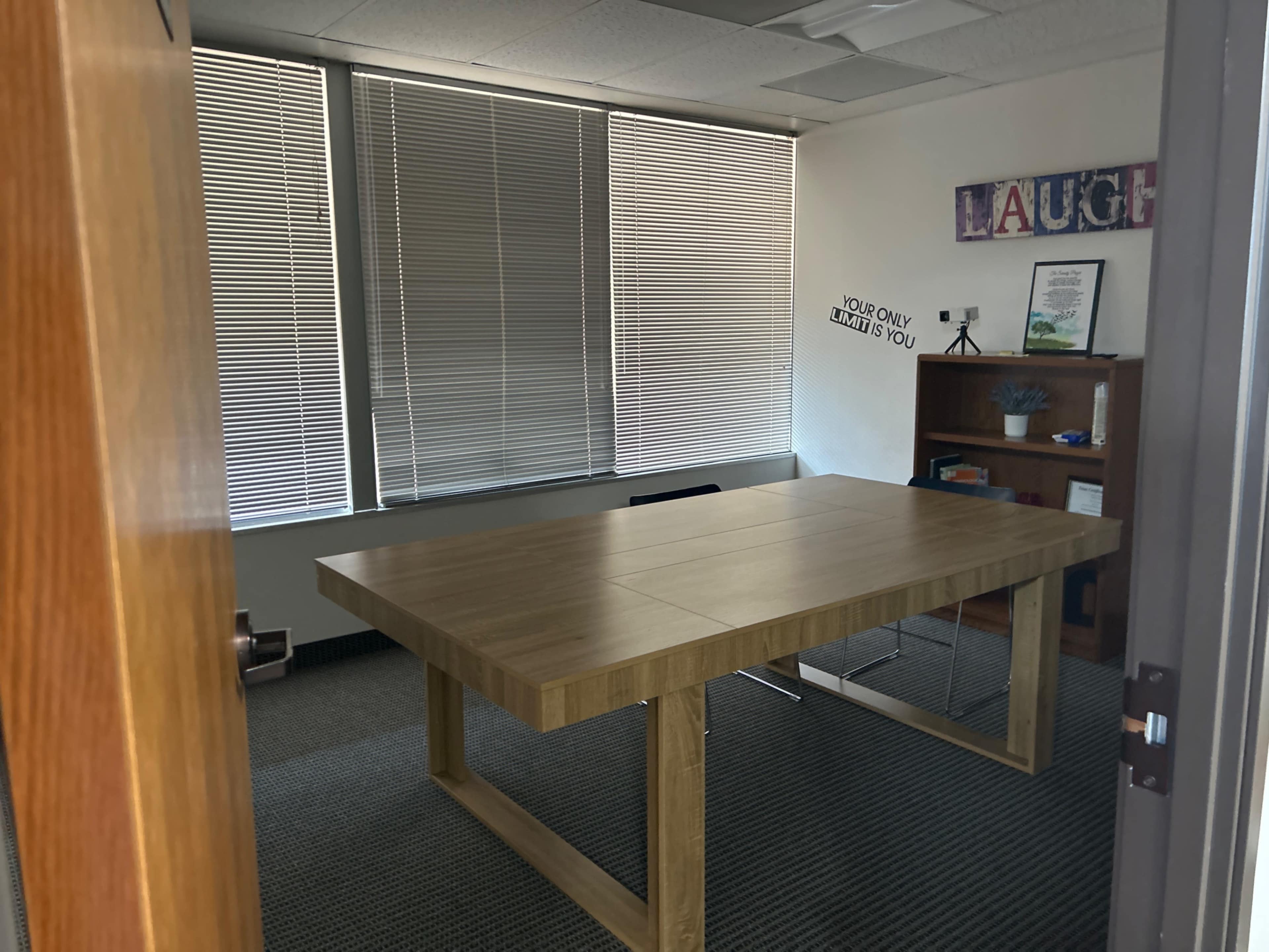 A wooden table and a chair are positioned in a sparsely furnished office with large windows covered by blinds.