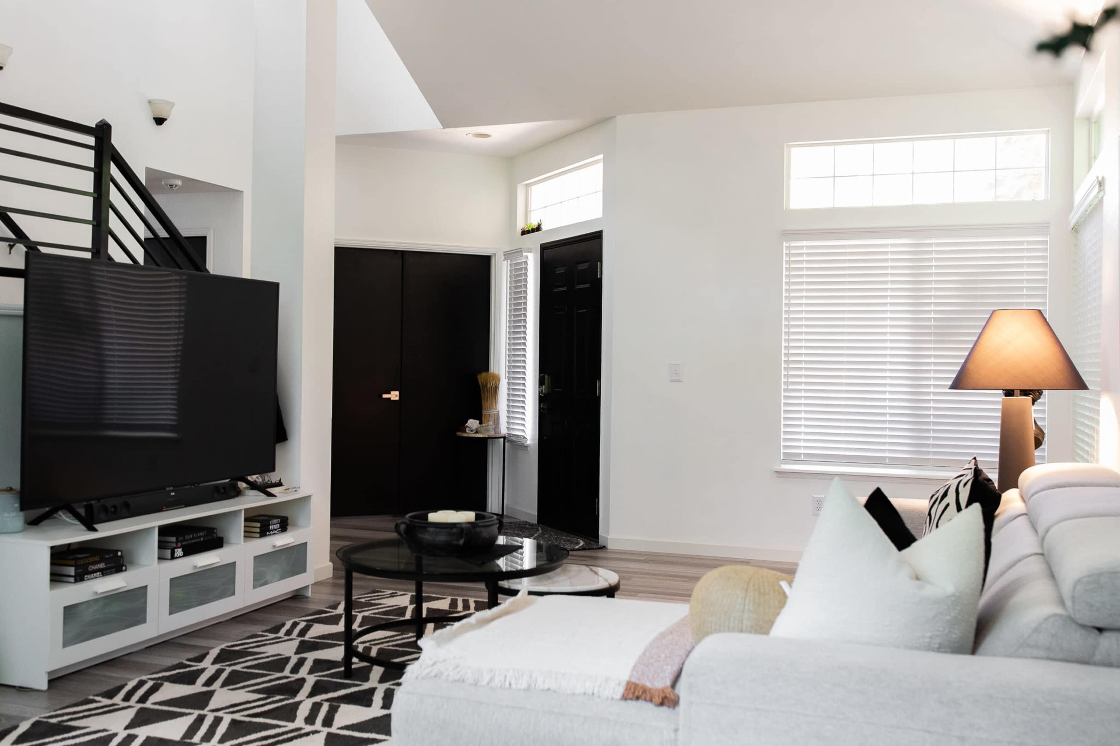 A modern living room featuring a light-colored sofa, a large television on a white stand, a circular coffee table, and large windows providing natural light.