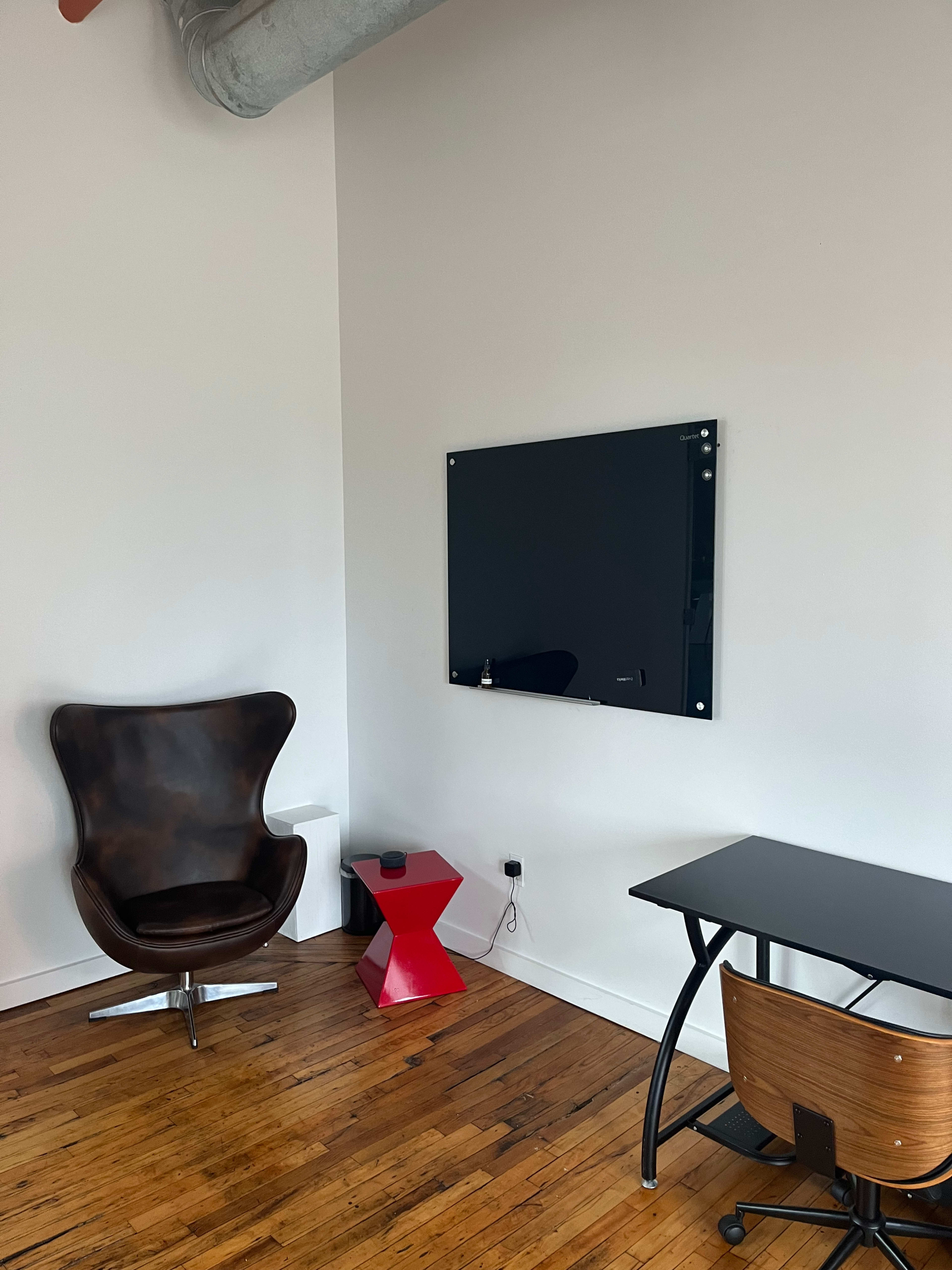 A modern, minimalist room featuring a brown chair, a red side table, a glass board mounted on the wall, and a black desk with a wooden chair.