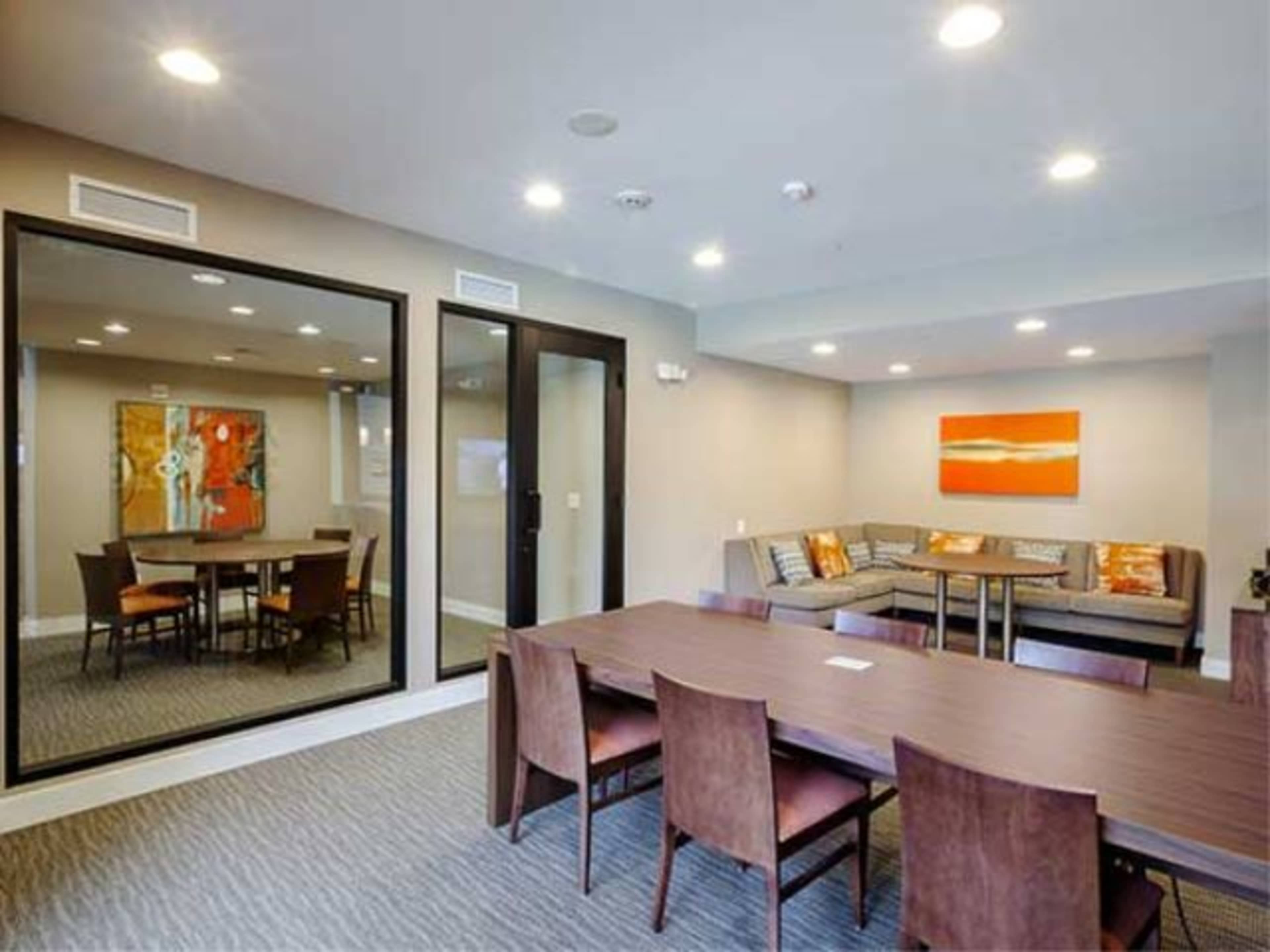 The room features a dining area with a large table surrounded by chairs, a lounge area with a sectional sofa, and a mirror reflecting artwork on the opposite wall.