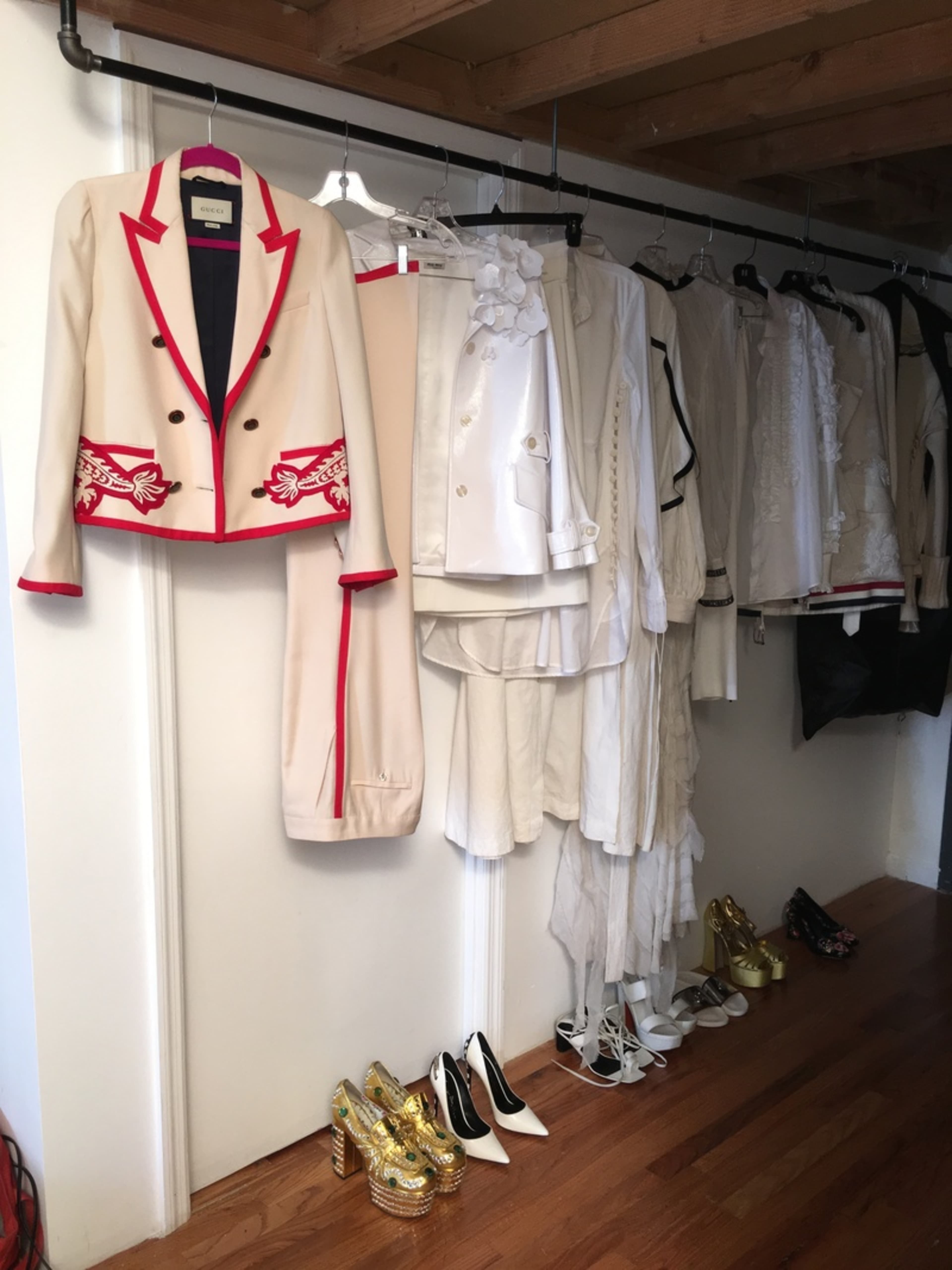 A variety of stylish clothing, including a beige suit with red detailing and several white garments, are hung on a rack along a wall, with shoes displayed on the floor below.