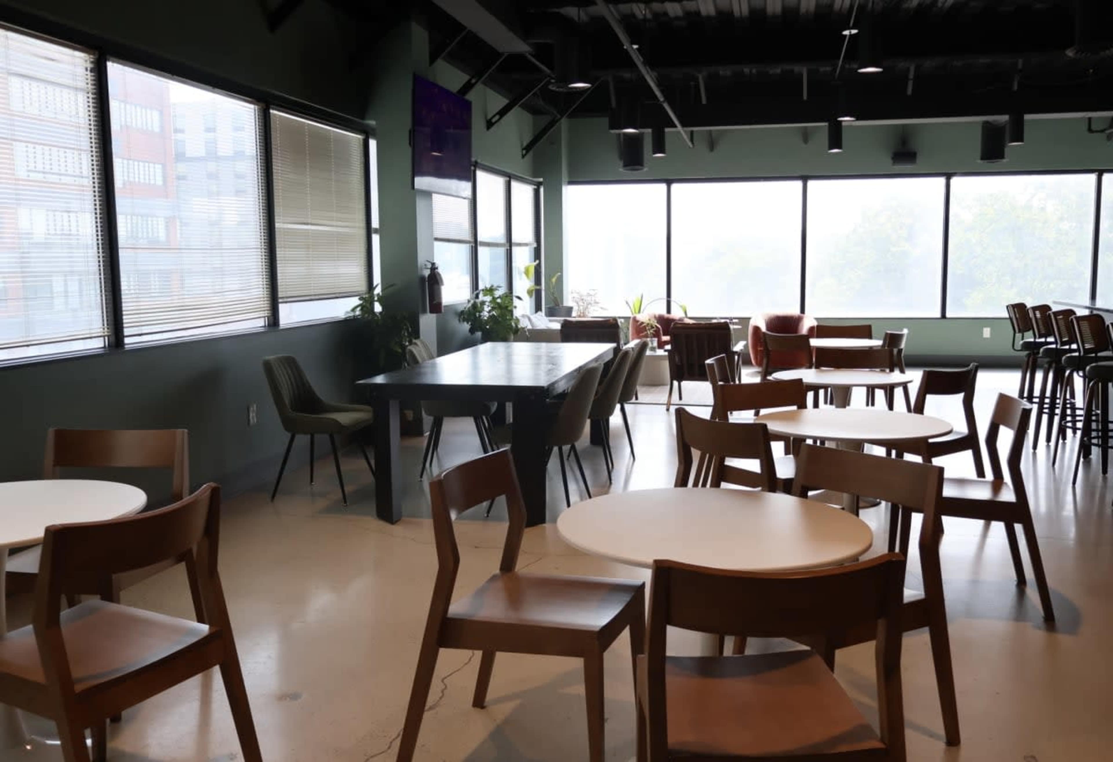 The image shows a spacious room with large windows, featuring several tables and chairs, along with plants and a bar area.