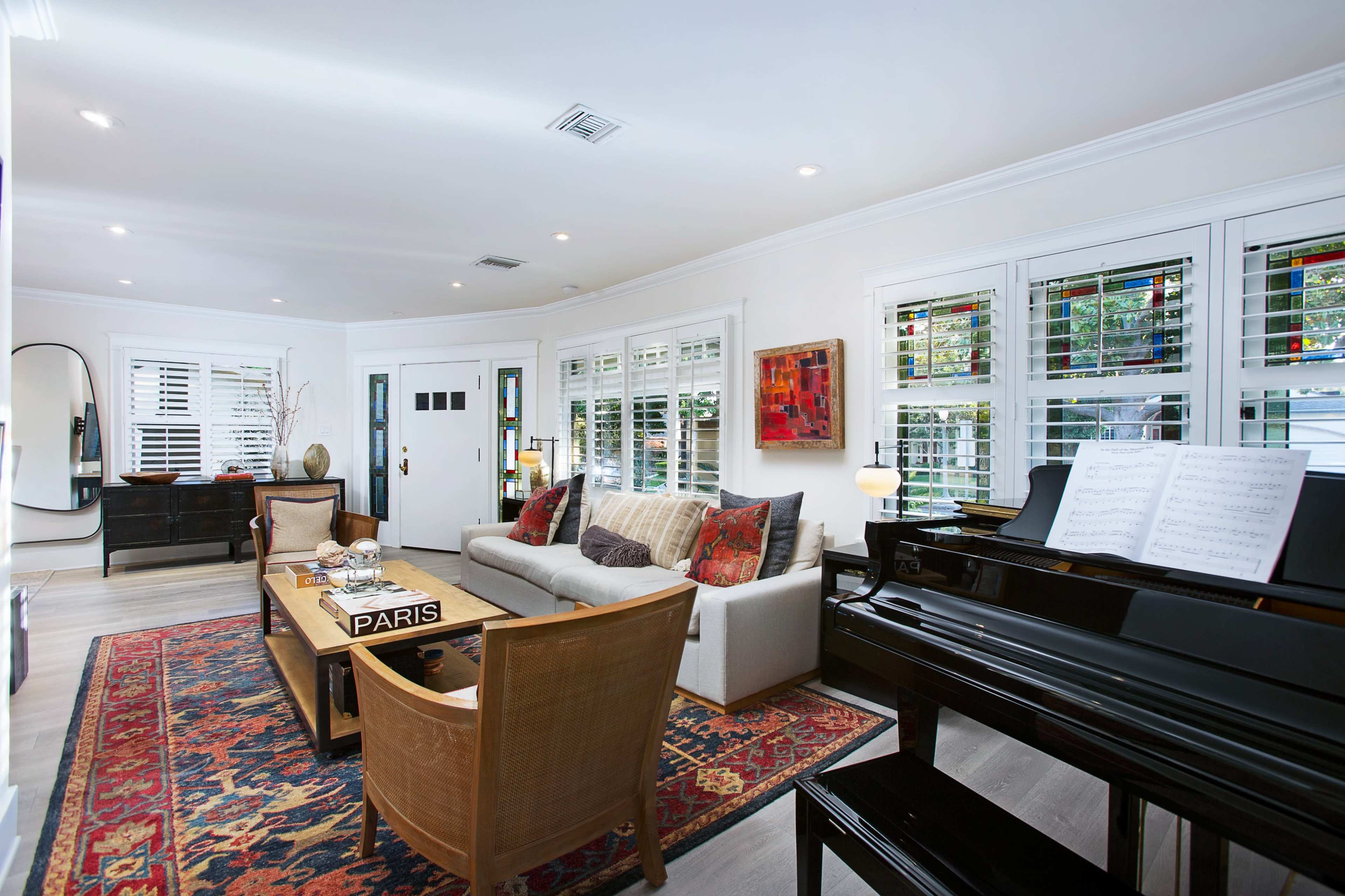 A bright living room featuring a piano, a comfortable sofa with patterned cushions, and a coffee table displaying the word "PARIS," all set against a backdrop of large windows with shutters.