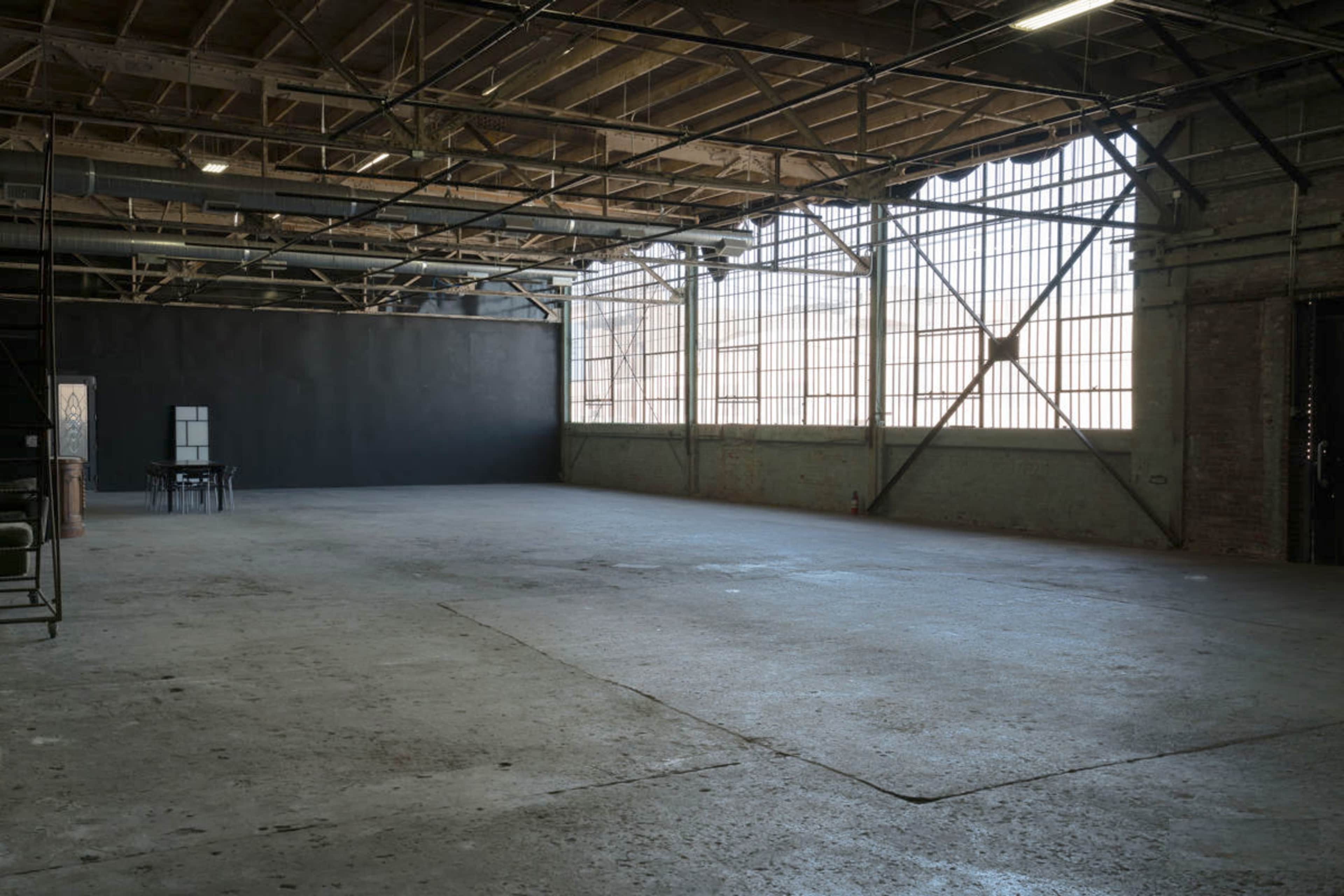 The image shows a spacious, empty industrial room with large windows and exposed beams.