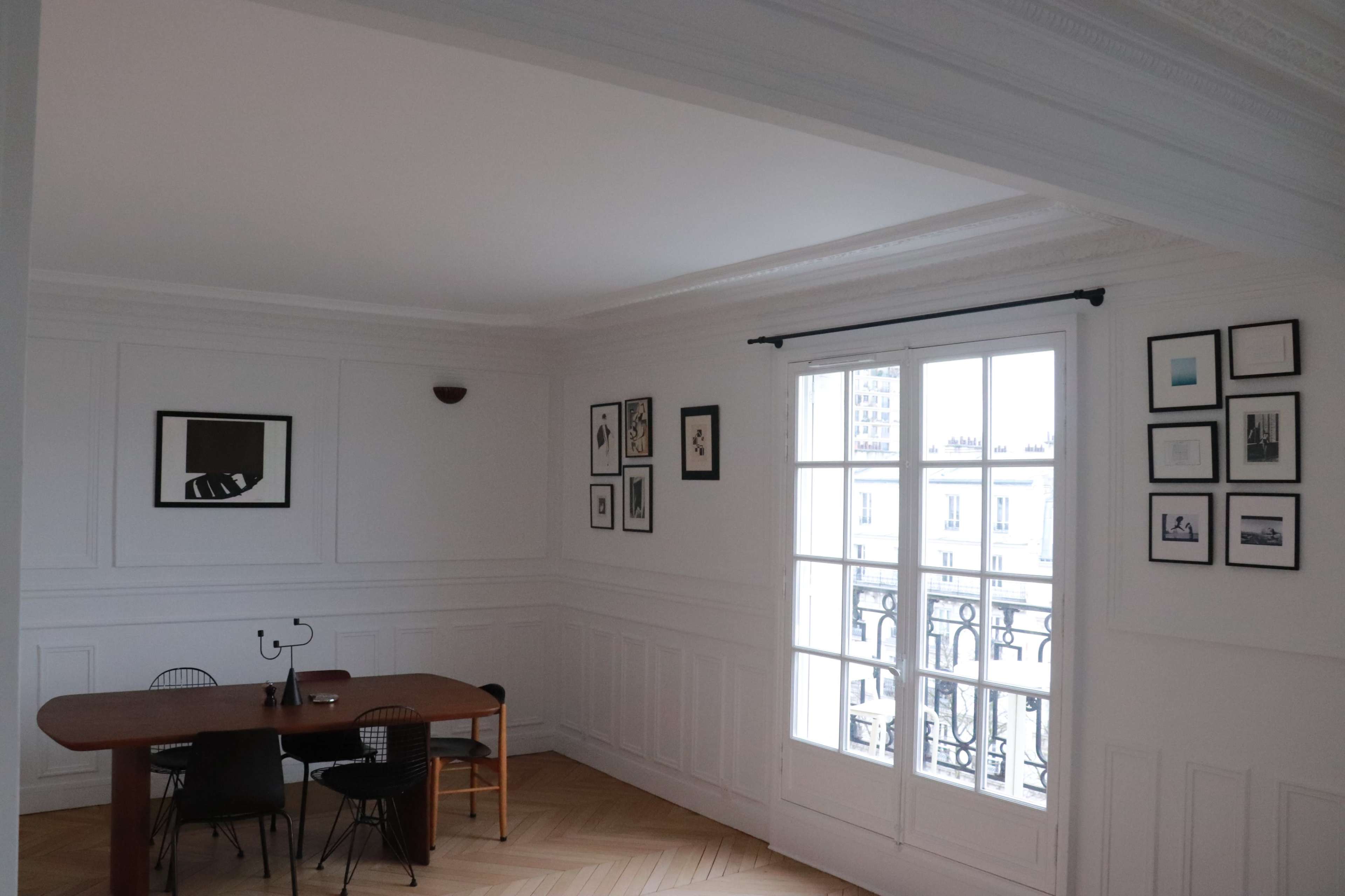 A bright, minimalist room features a wooden dining table surrounded by black chairs and walls adorned with framed artwork.