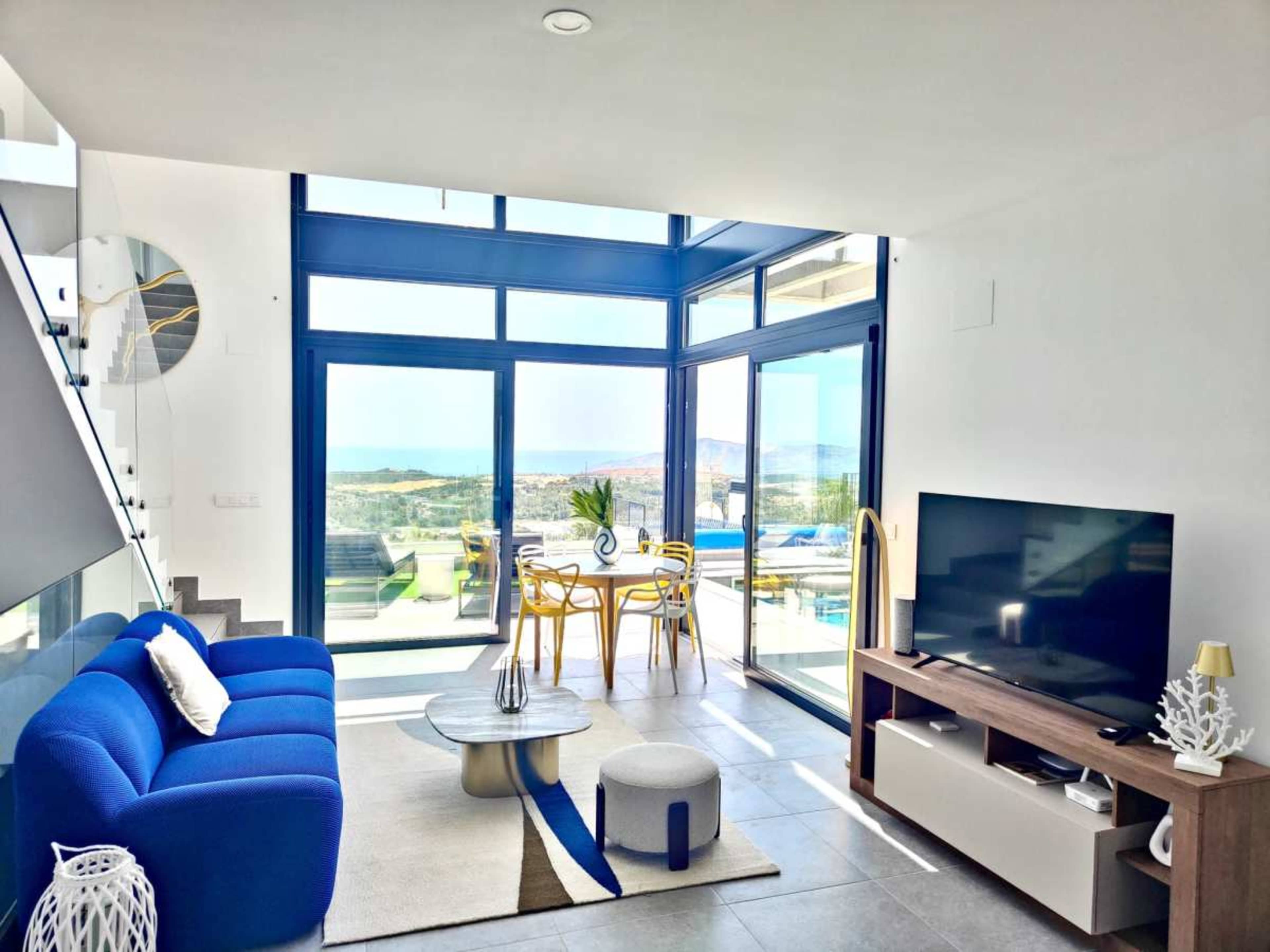 A modern living room features a blue sofa, a television, and floor-to-ceiling windows that overlook a scenic view.