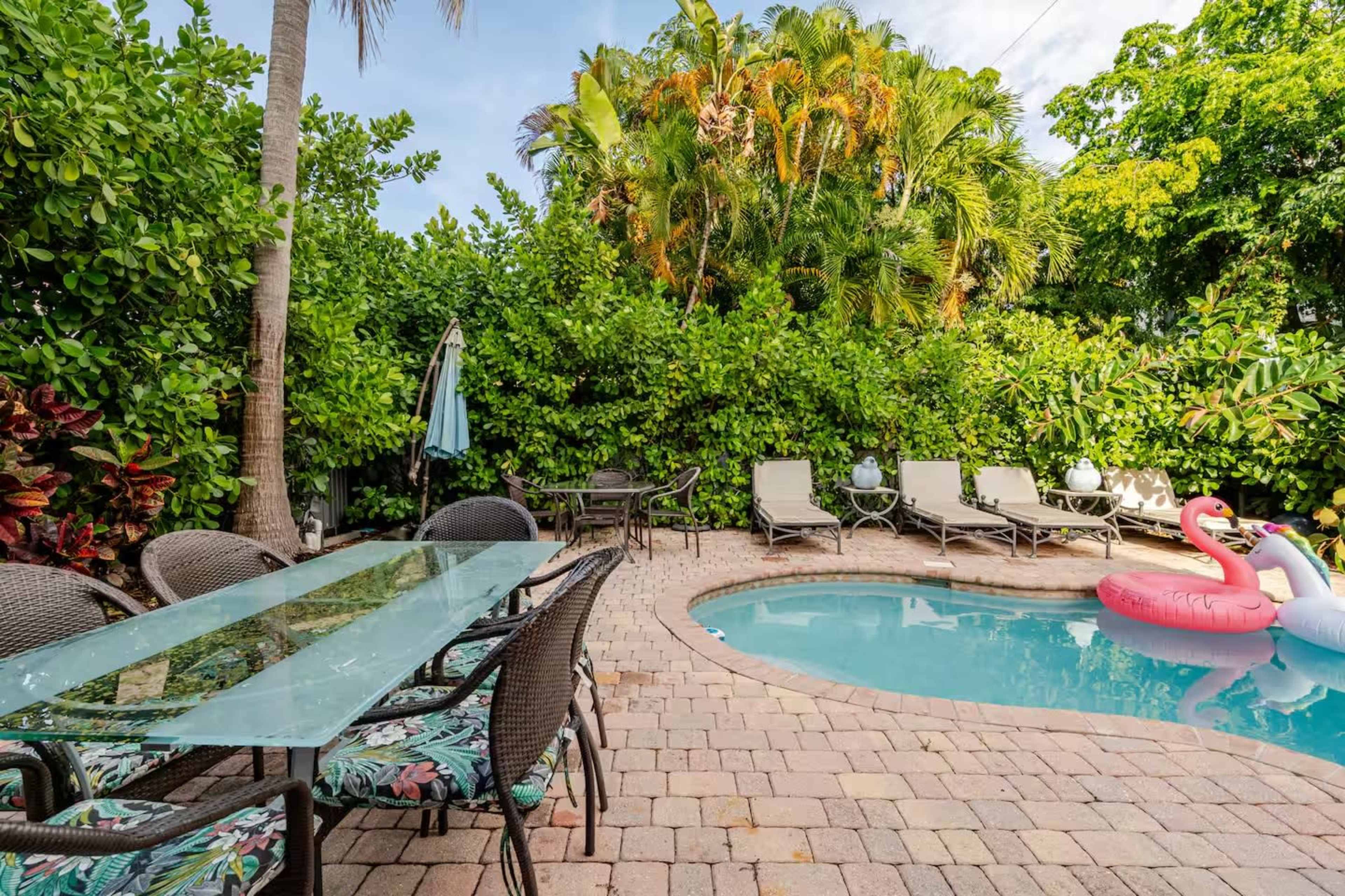 A stone patio surrounds a small pool with lounge chairs and a dining table, set against a backdrop of lush greenery.