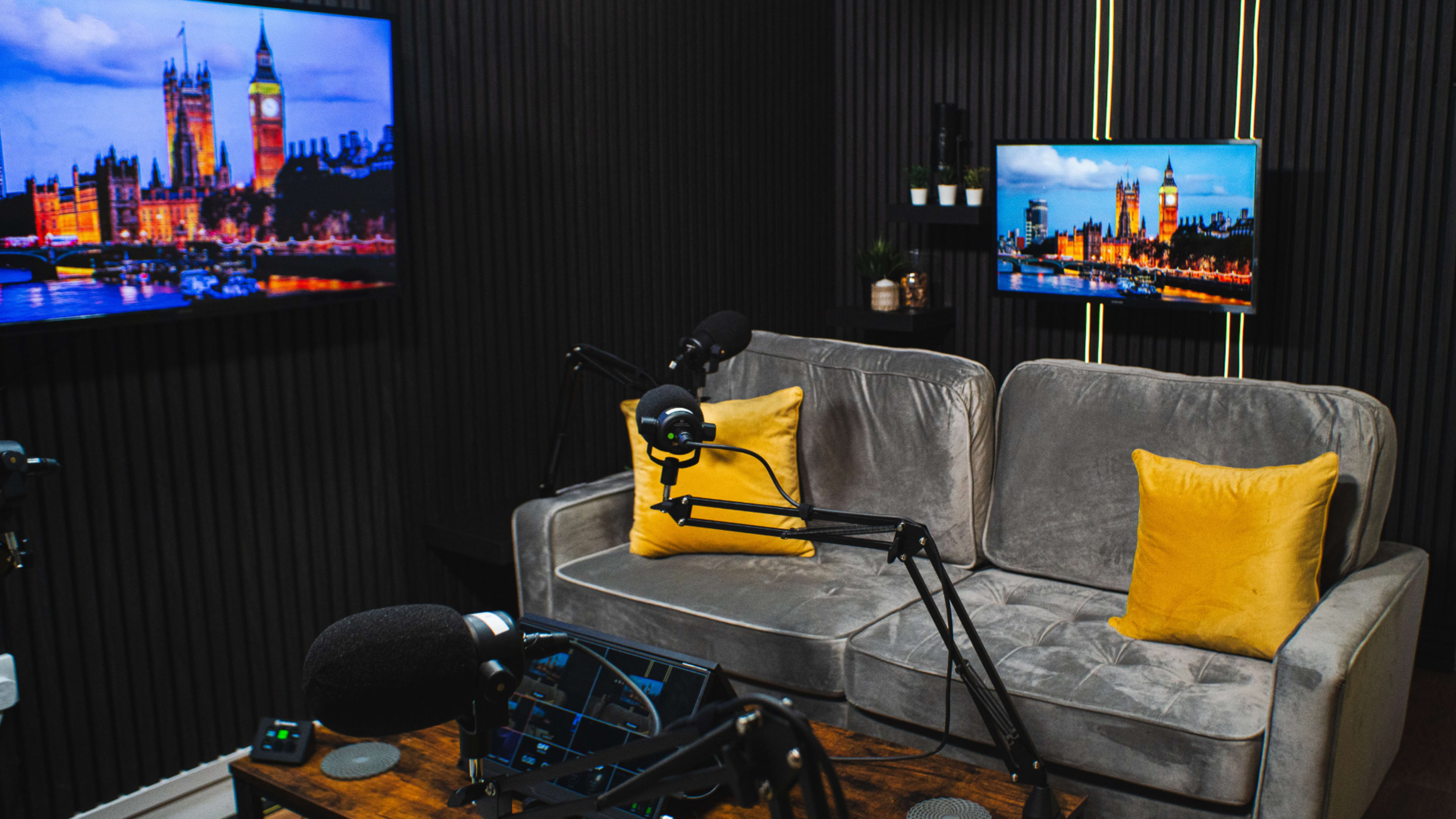 A podcasting studio with a gray couch, yellow pillows, and two screens displaying a view of a cityscape.