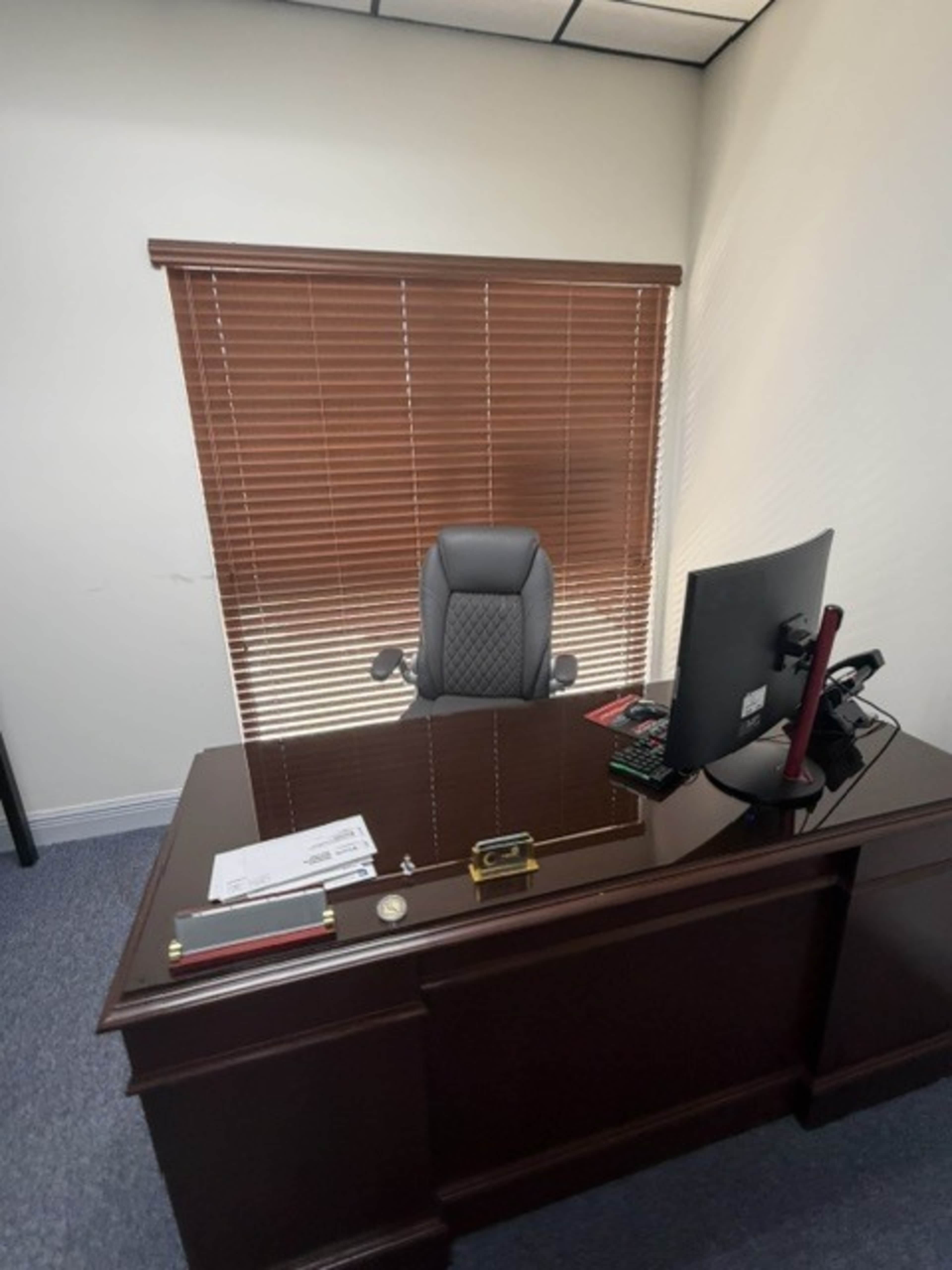 The image shows a modern office space with a wooden desk, a swivel chair, and a computer monitor facing a window with blinds.