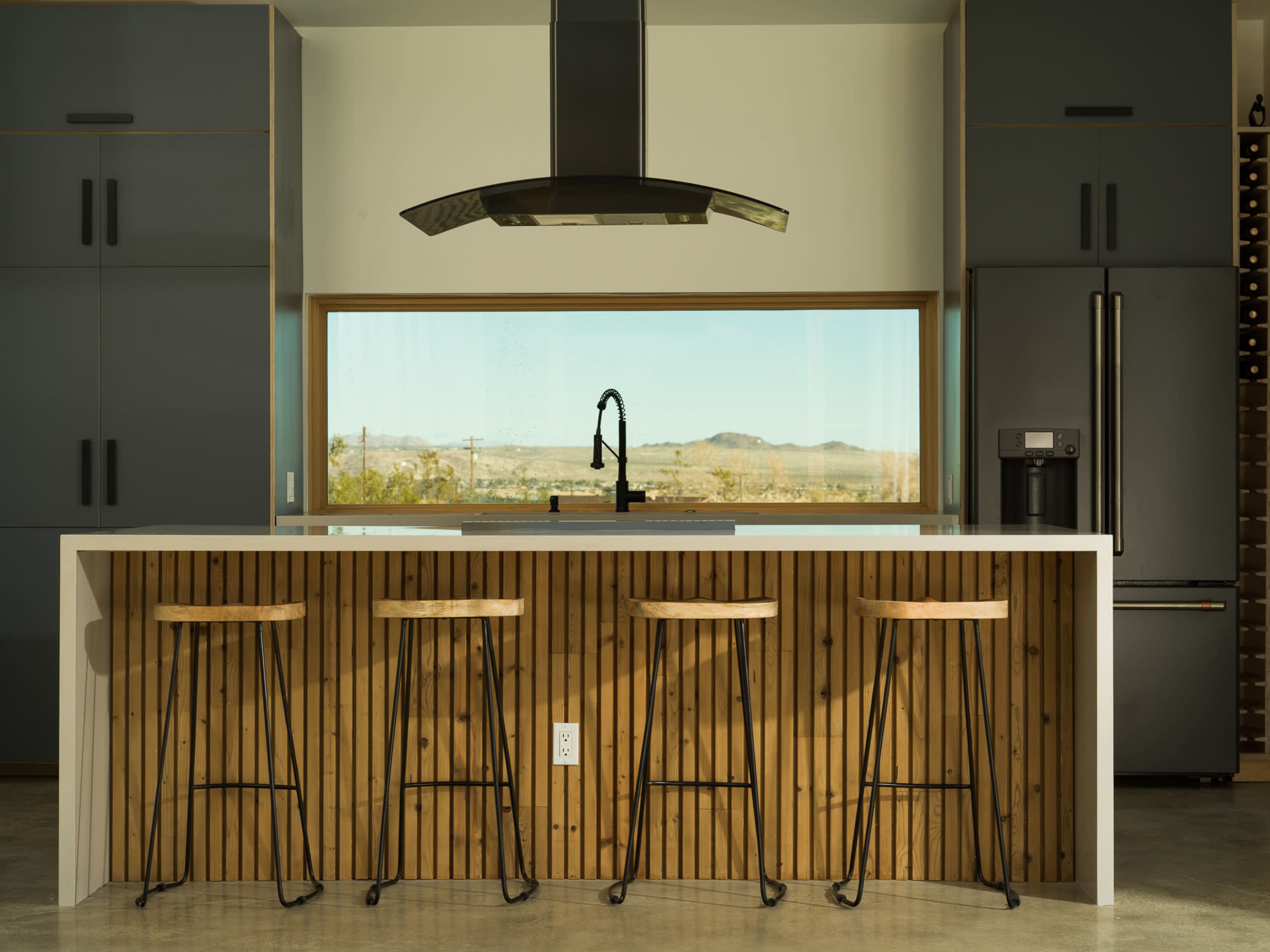 A modern kitchen features a central island with four bar stools, a sleek hood above the cooktop, and large windows offering a view of the landscape outside.