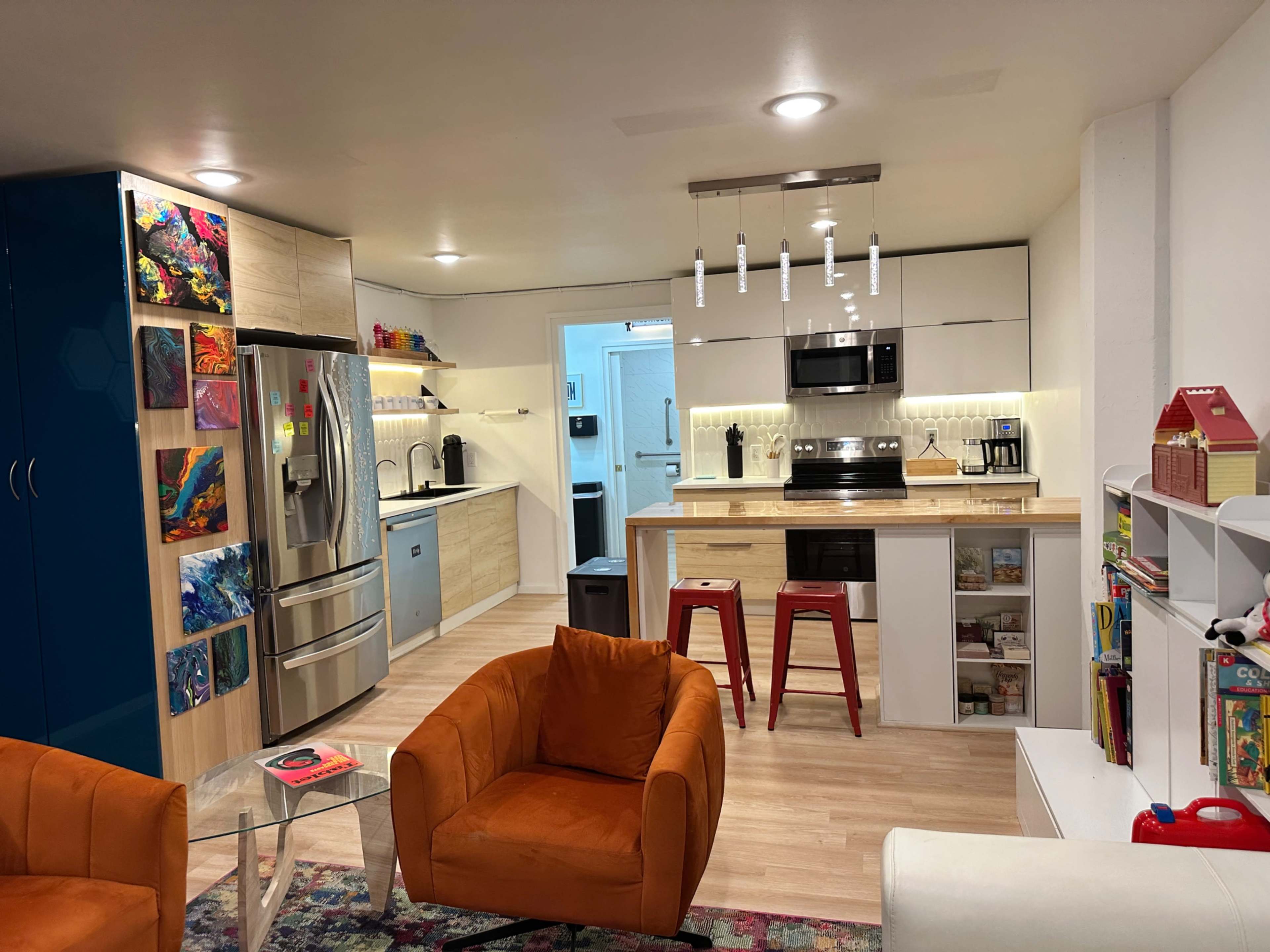 The image shows a modern kitchen and living area featuring a stainless steel refrigerator, a white kitchen island with bar stools, and orange accent chairs.