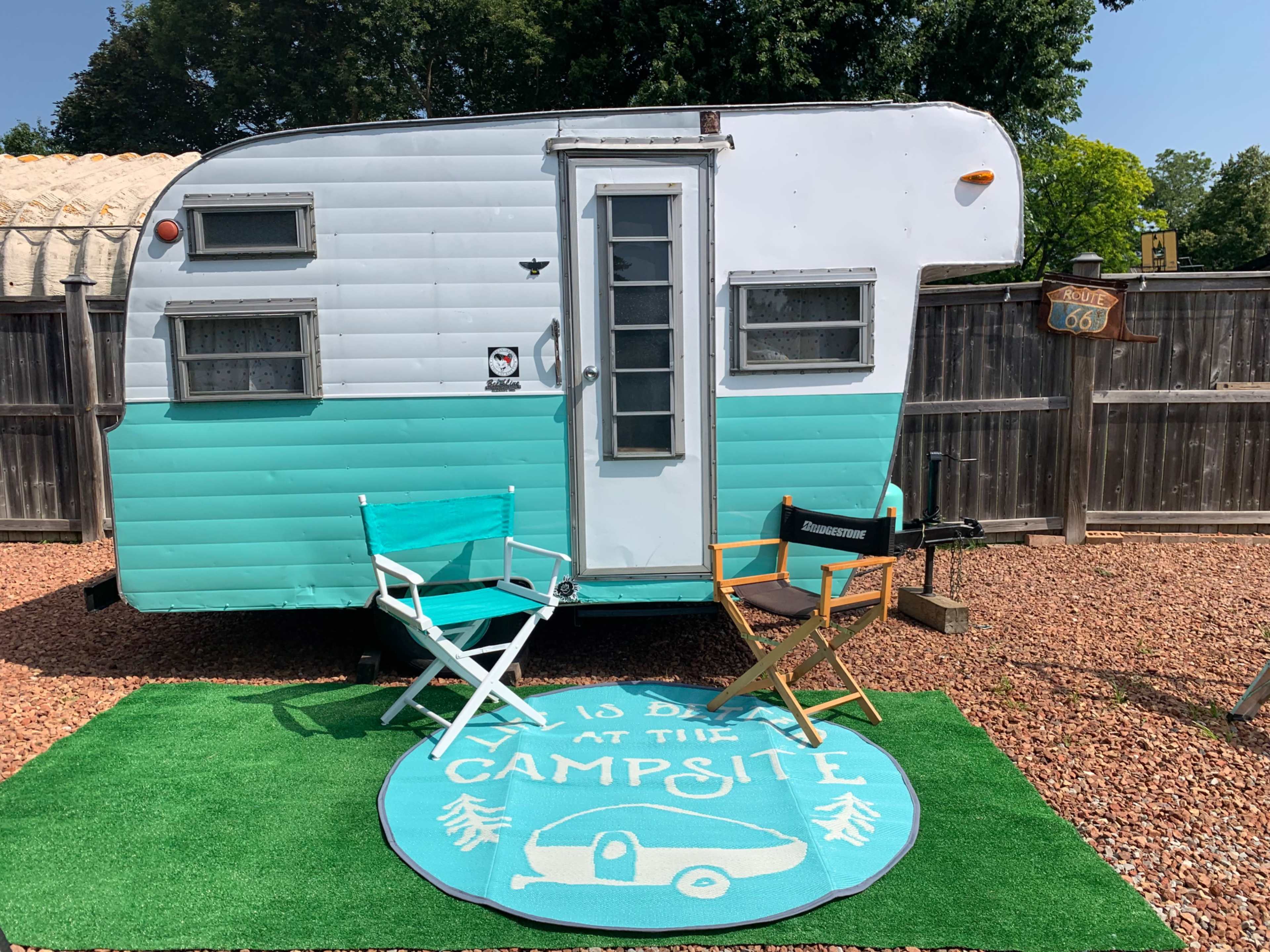 A vintage camper is parked in a gravel area with two folding chairs and a green doormat that reads "It's a Day at the Campsite."