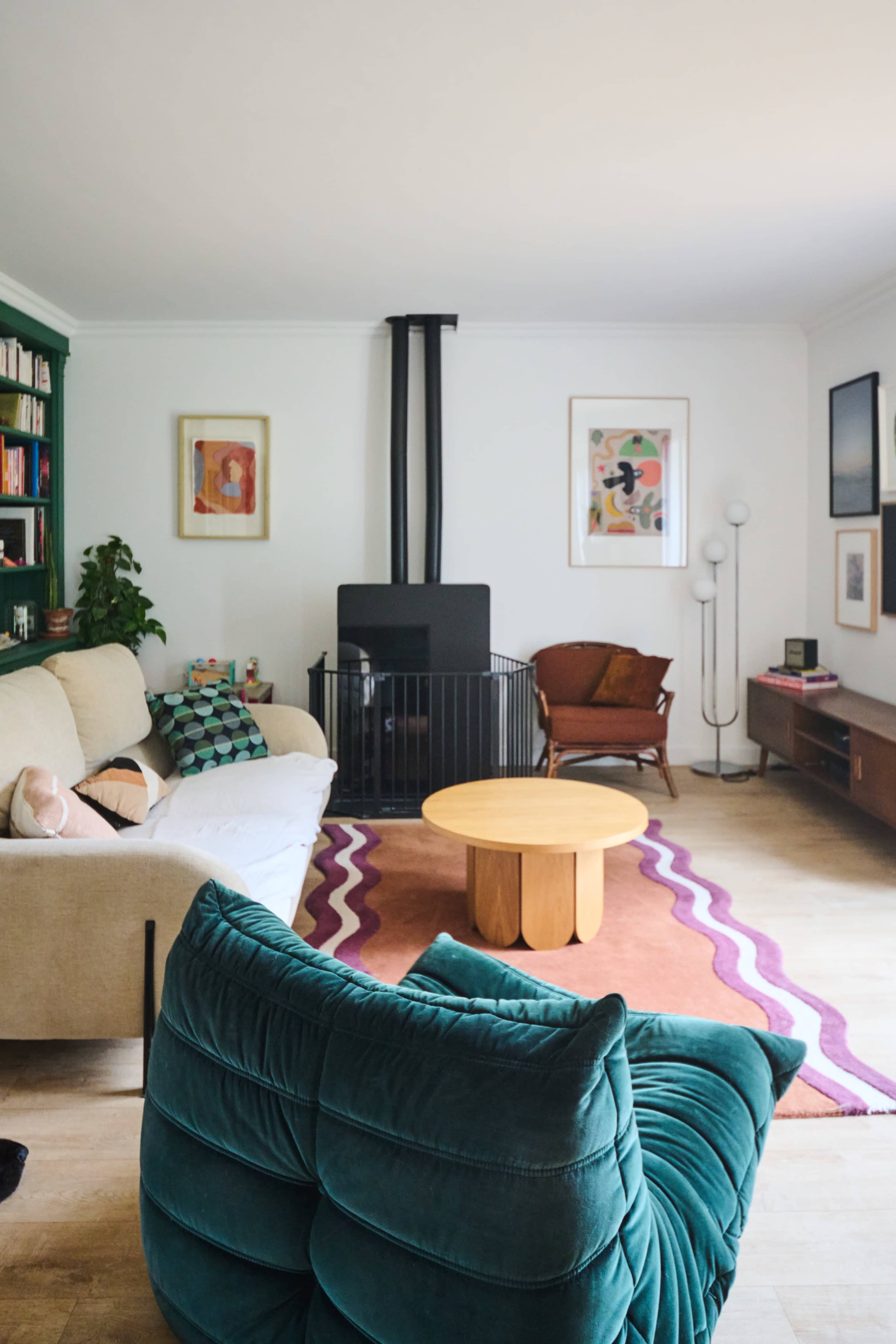 A cozy living room features a green sofa, a circular wooden coffee table, a fireplace, and various artworks on the walls.