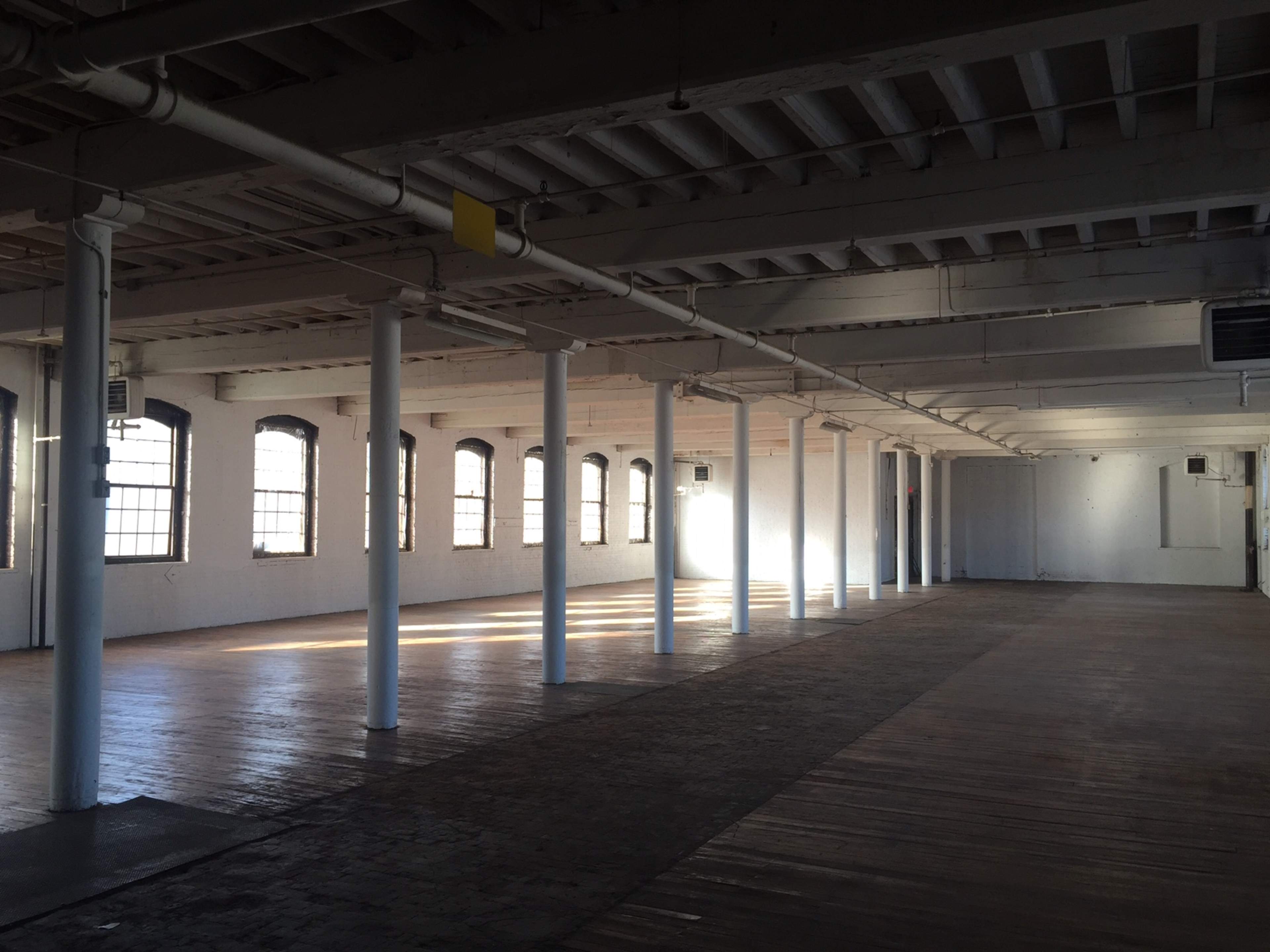 A spacious, empty room with wooden flooring and rows of white columns, illuminated by sunlight filtering through large windows.
