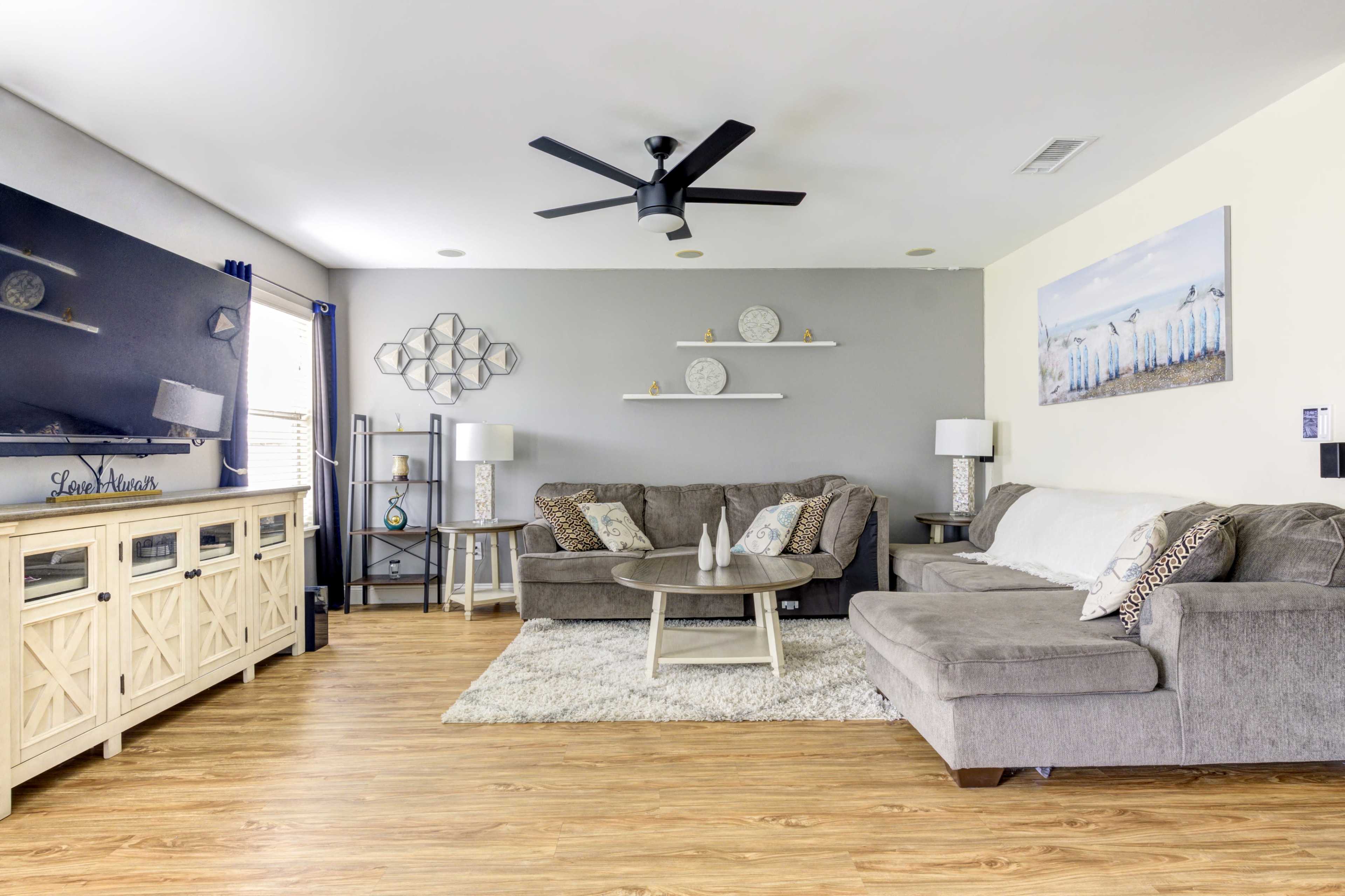 The living room features a gray sofa and loveseat arranged around a circular coffee table, with a television and decorative shelves on the wall.