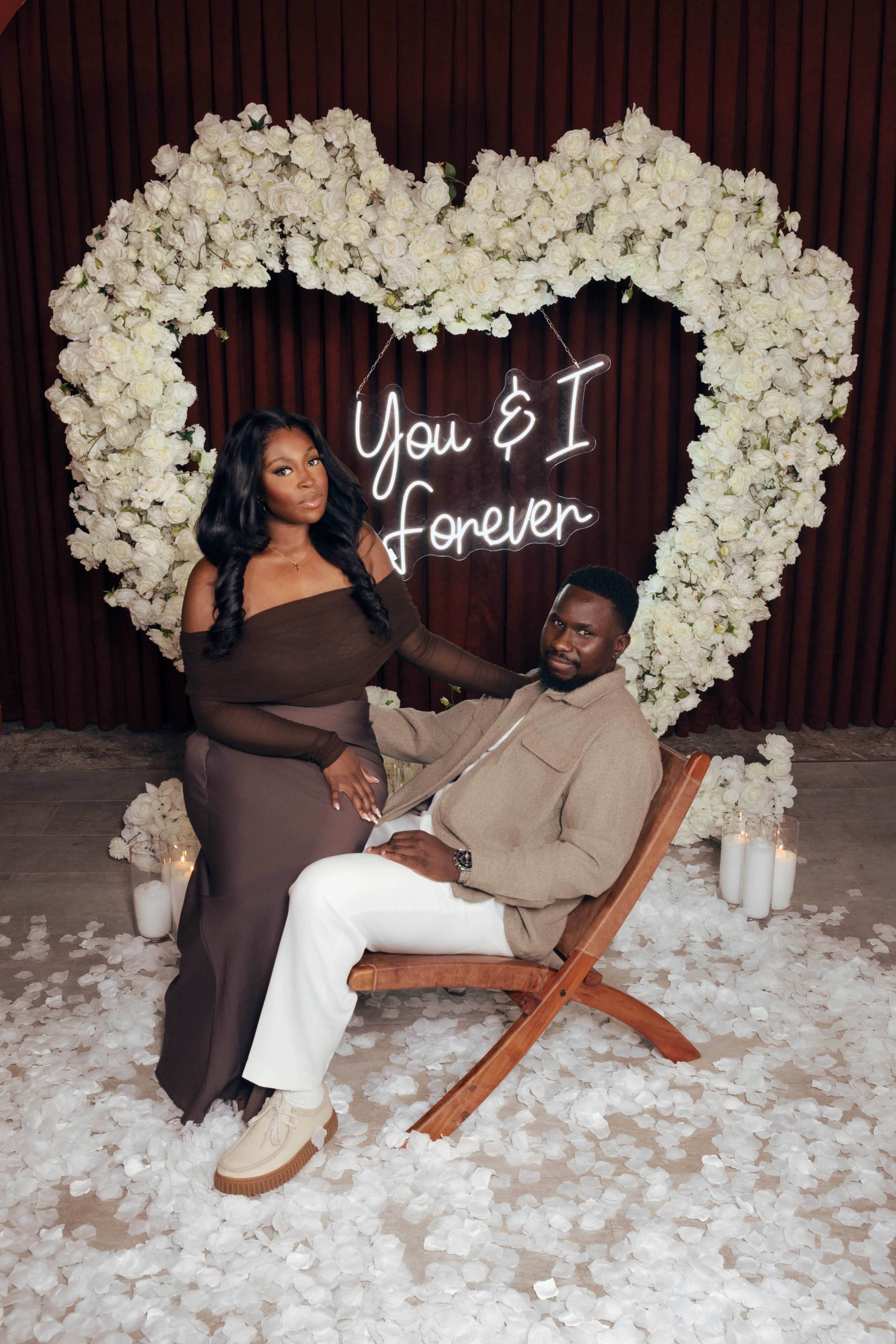A couple poses in front of a heart-shaped floral arrangement and a neon sign that reads "You & I Forever."