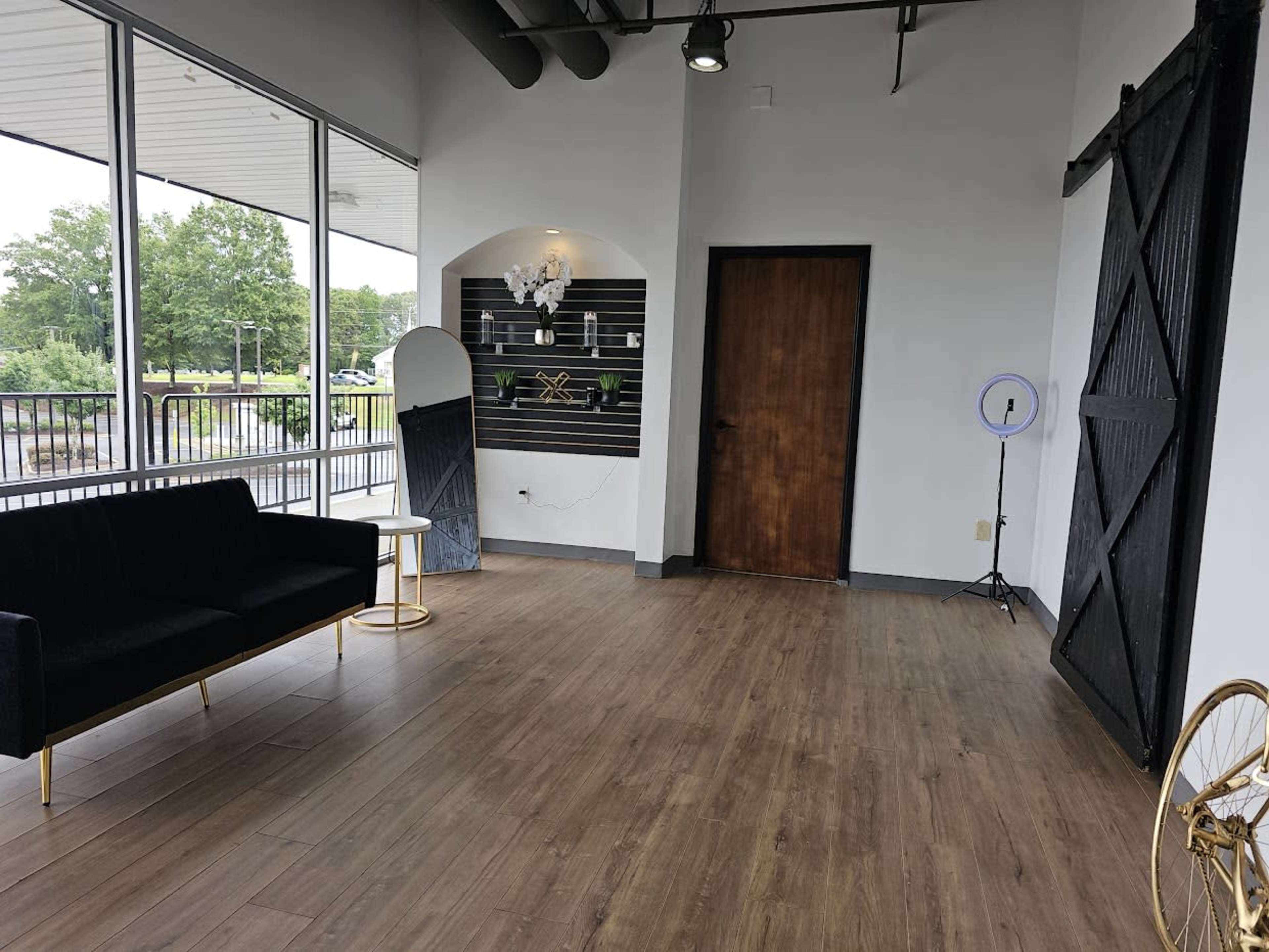 An interior space featuring a black sofa, a small round table, a large mirror, and a wooden door, with large windows letting in natural light.