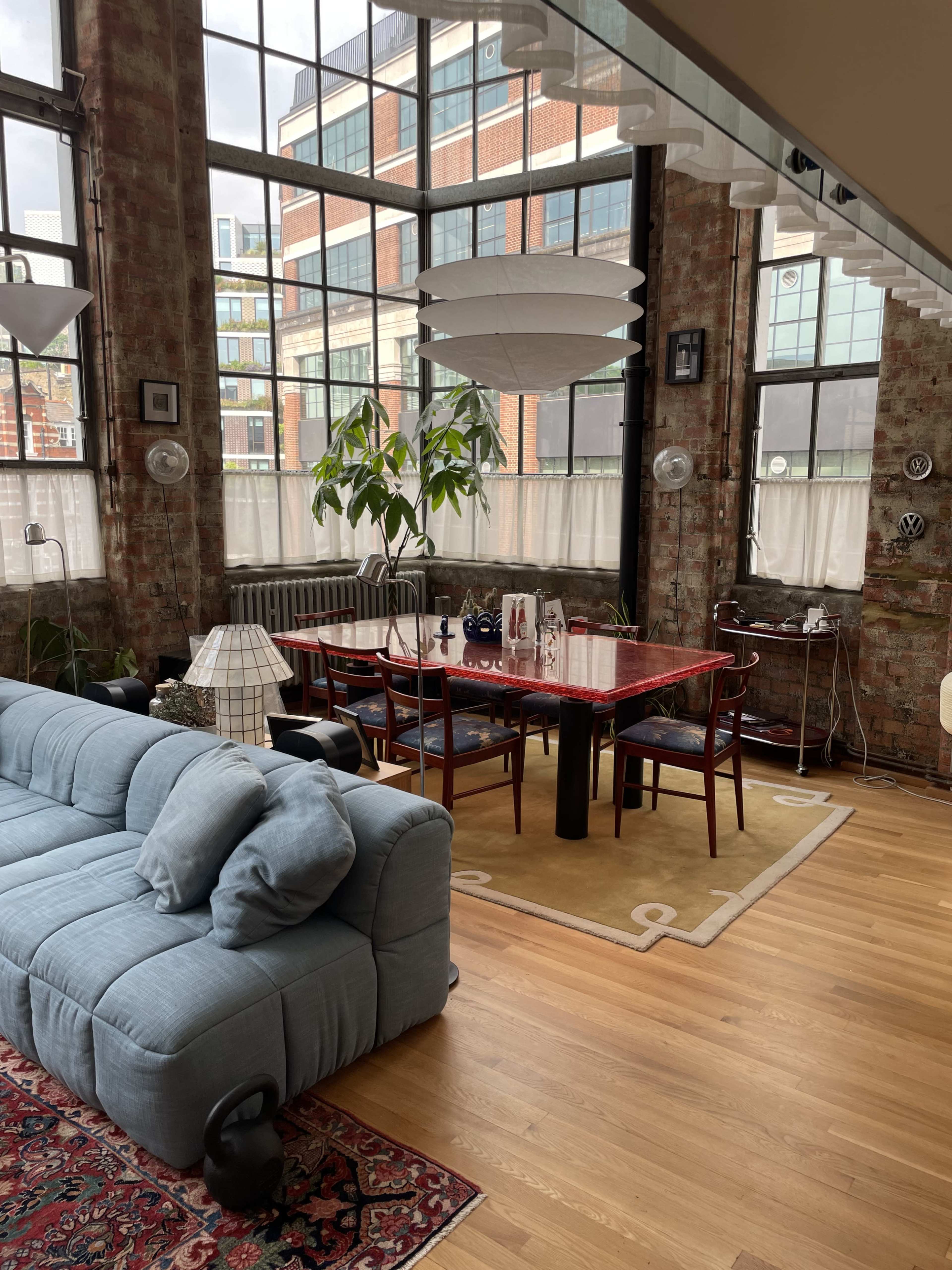 The image shows a modern dining area with a large table surrounded by chairs, a blue sofa, and large windows letting in natural light.