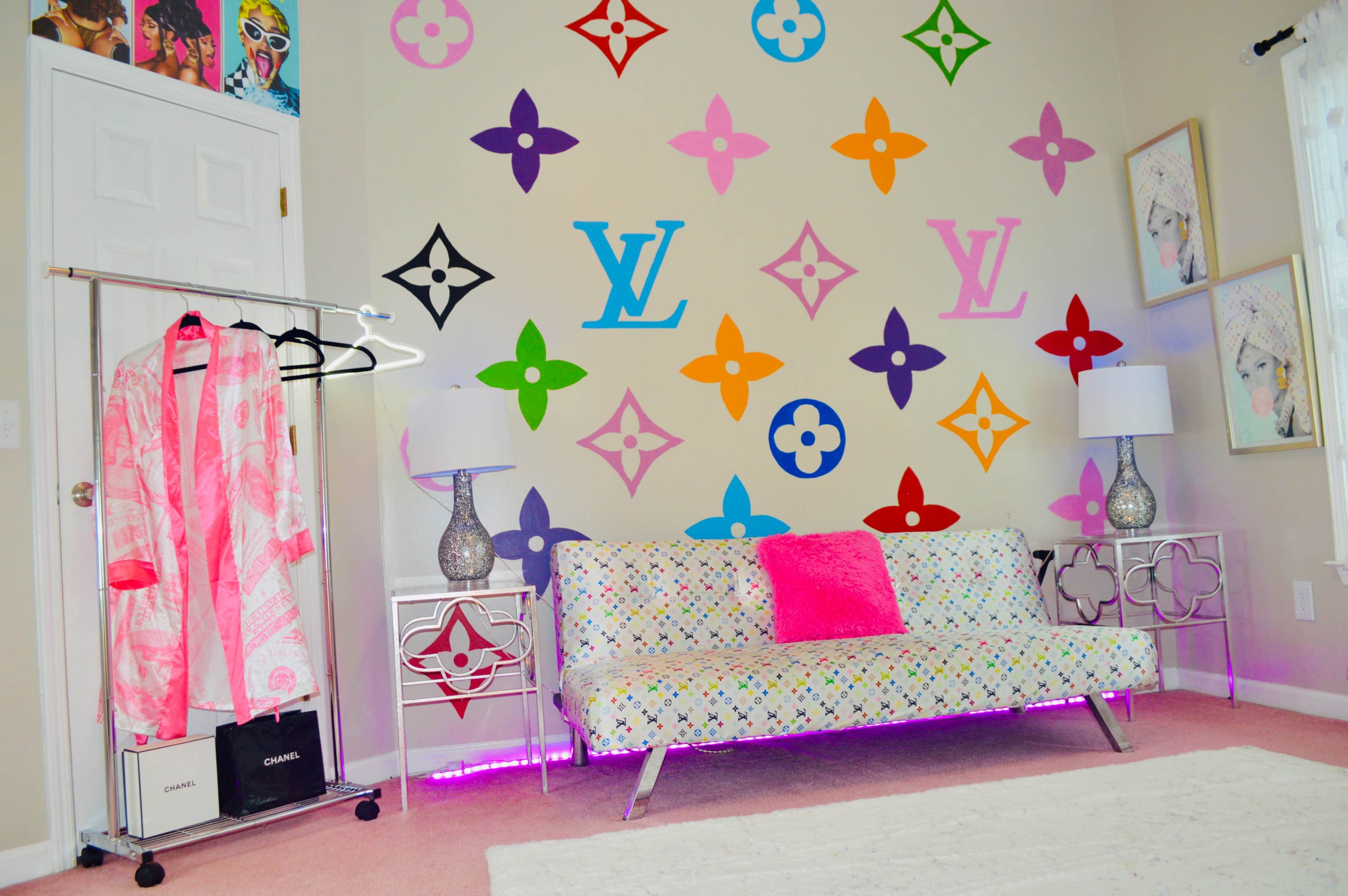 The image shows a brightly decorated room featuring a couch with a colorful pattern, a wall adorned with multicolored Louis Vuitton motifs, and a clothing rack beside it.