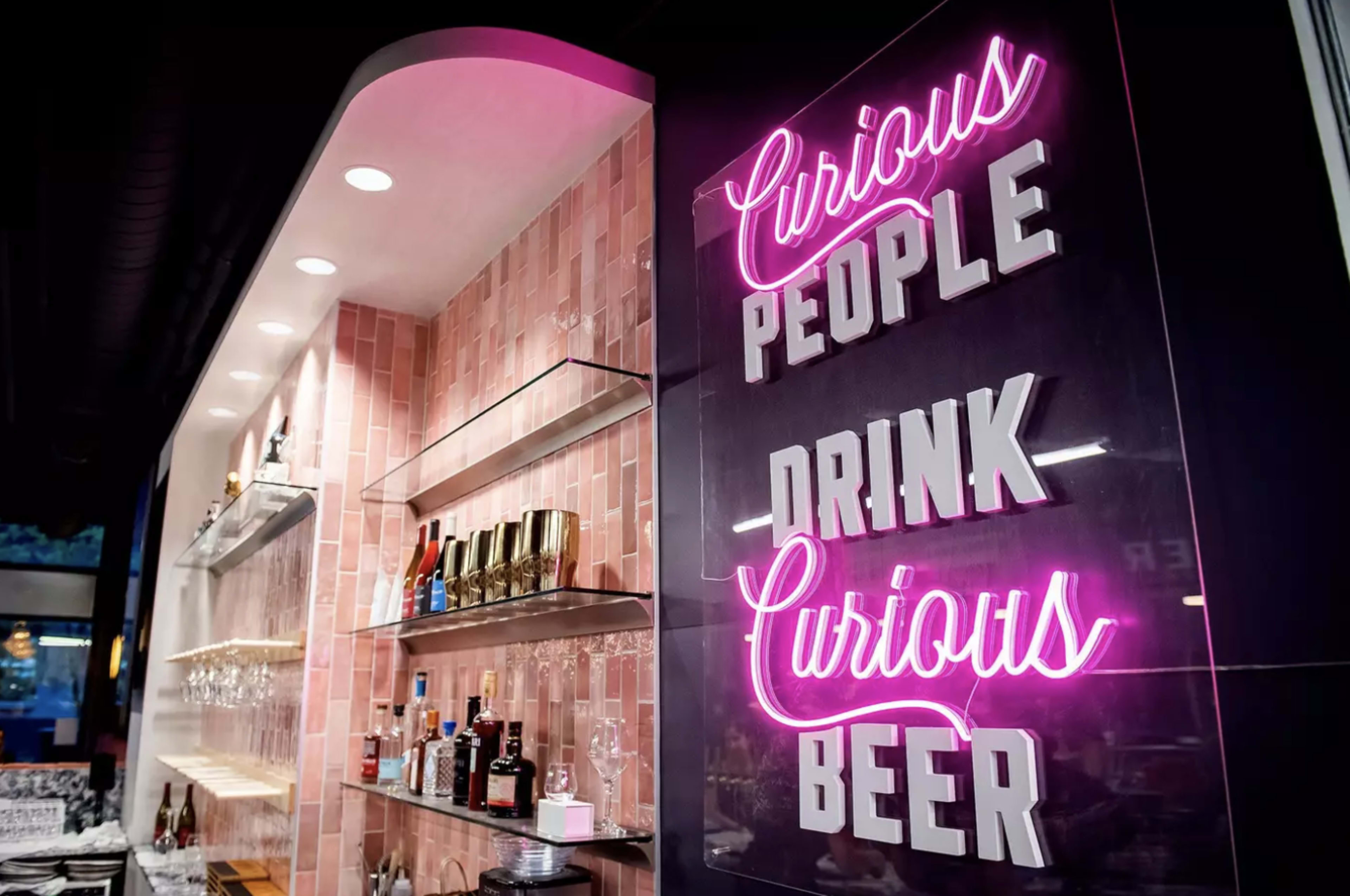 A bar area with a wall featuring a neon sign that reads "Curious People Drink Curious Beer," alongside shelves stocked with various bottles and glasses.