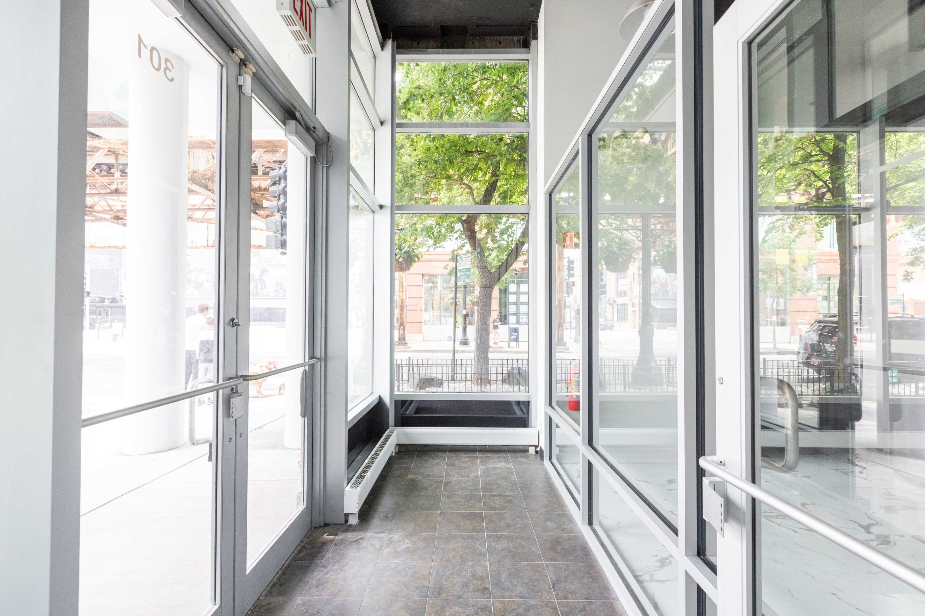 The image shows a well-lit corridor with large glass windows and doors, leading to a street view outside.