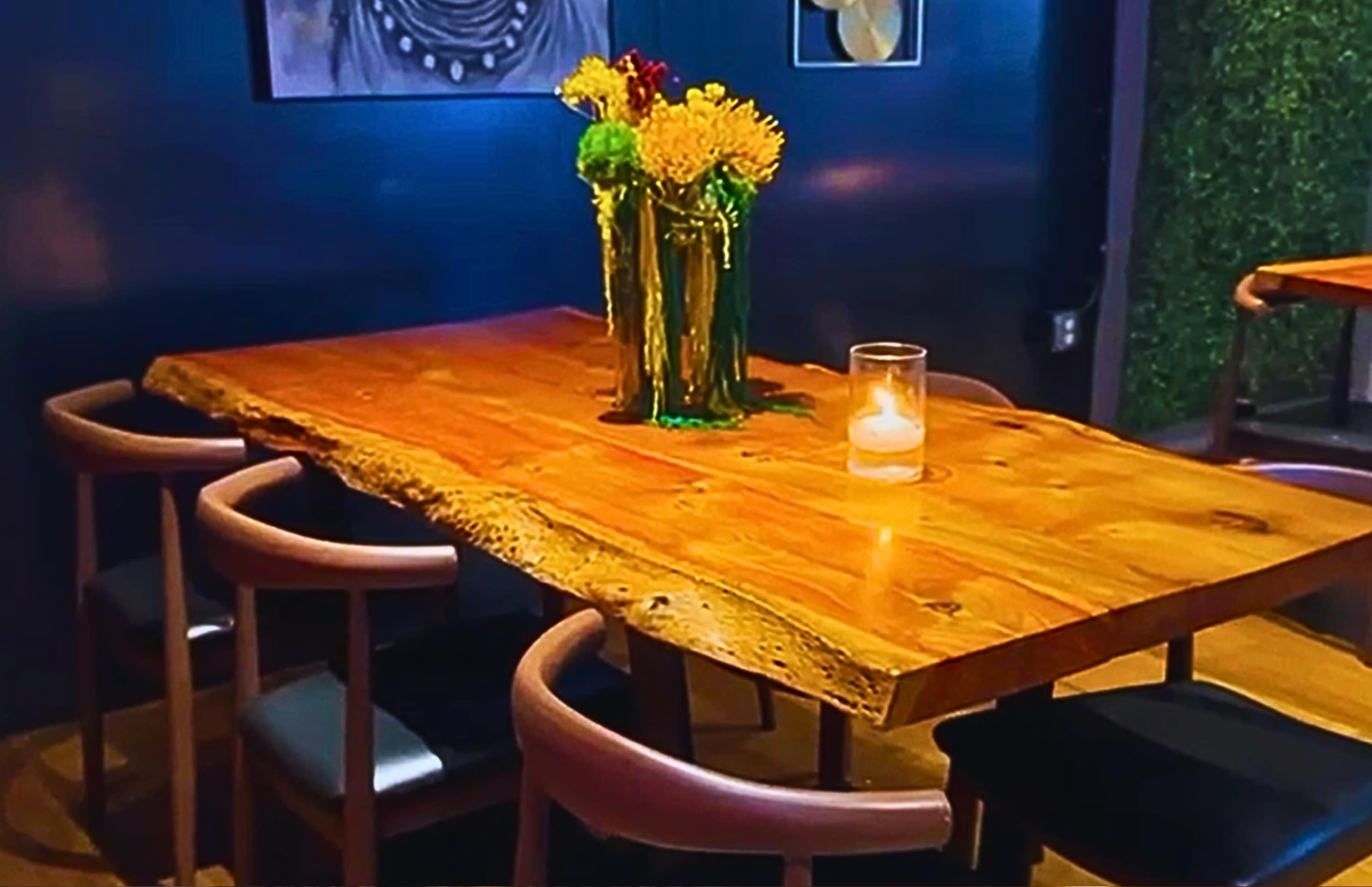 A wooden table with a natural edge is set with a candle and a vase of flowers, surrounded by four chairs in a dimly lit restaurant.