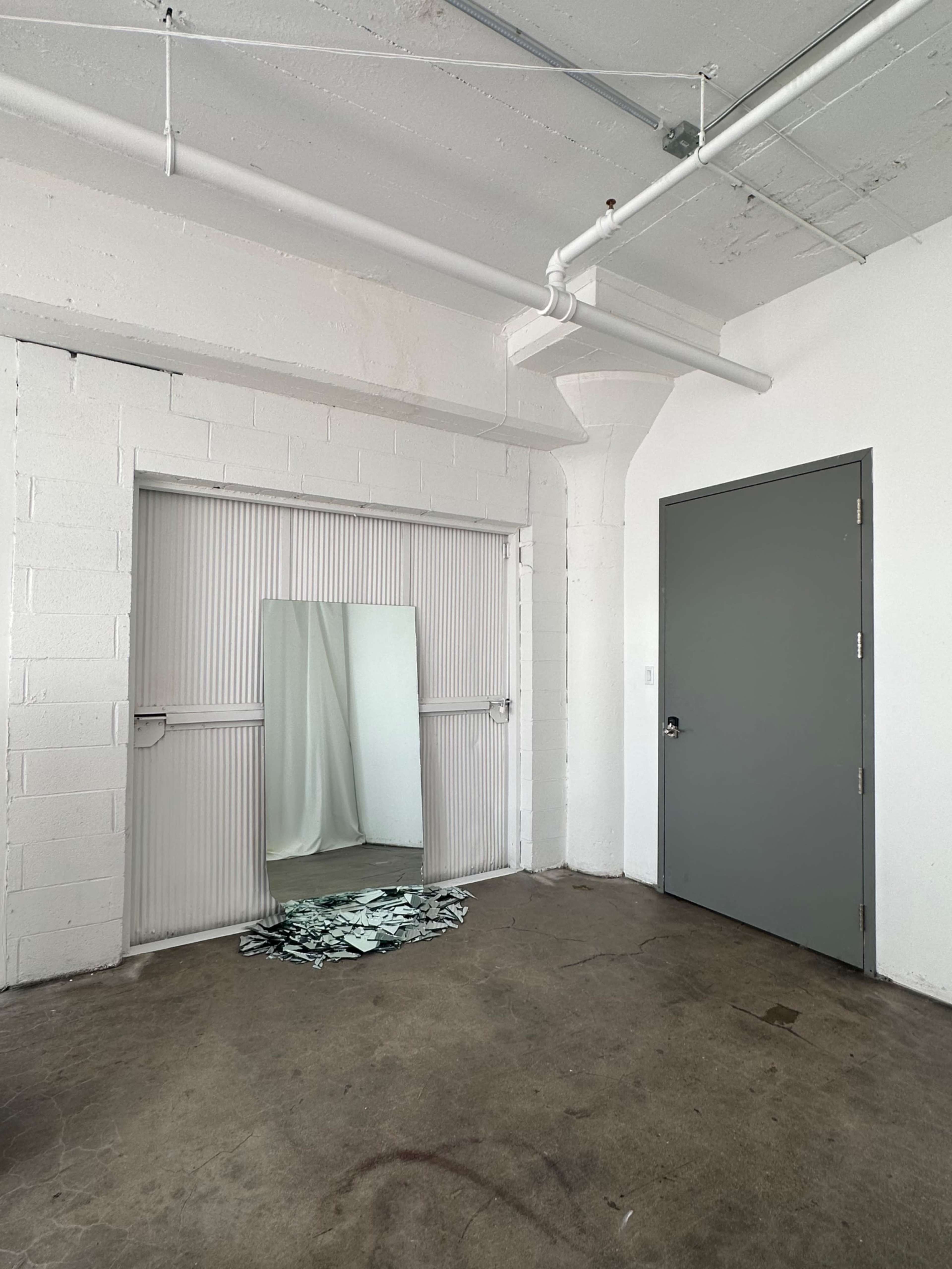 A large mirror leans against a wall in a sparse, industrial-style room with concrete flooring and a closed door in the corner.