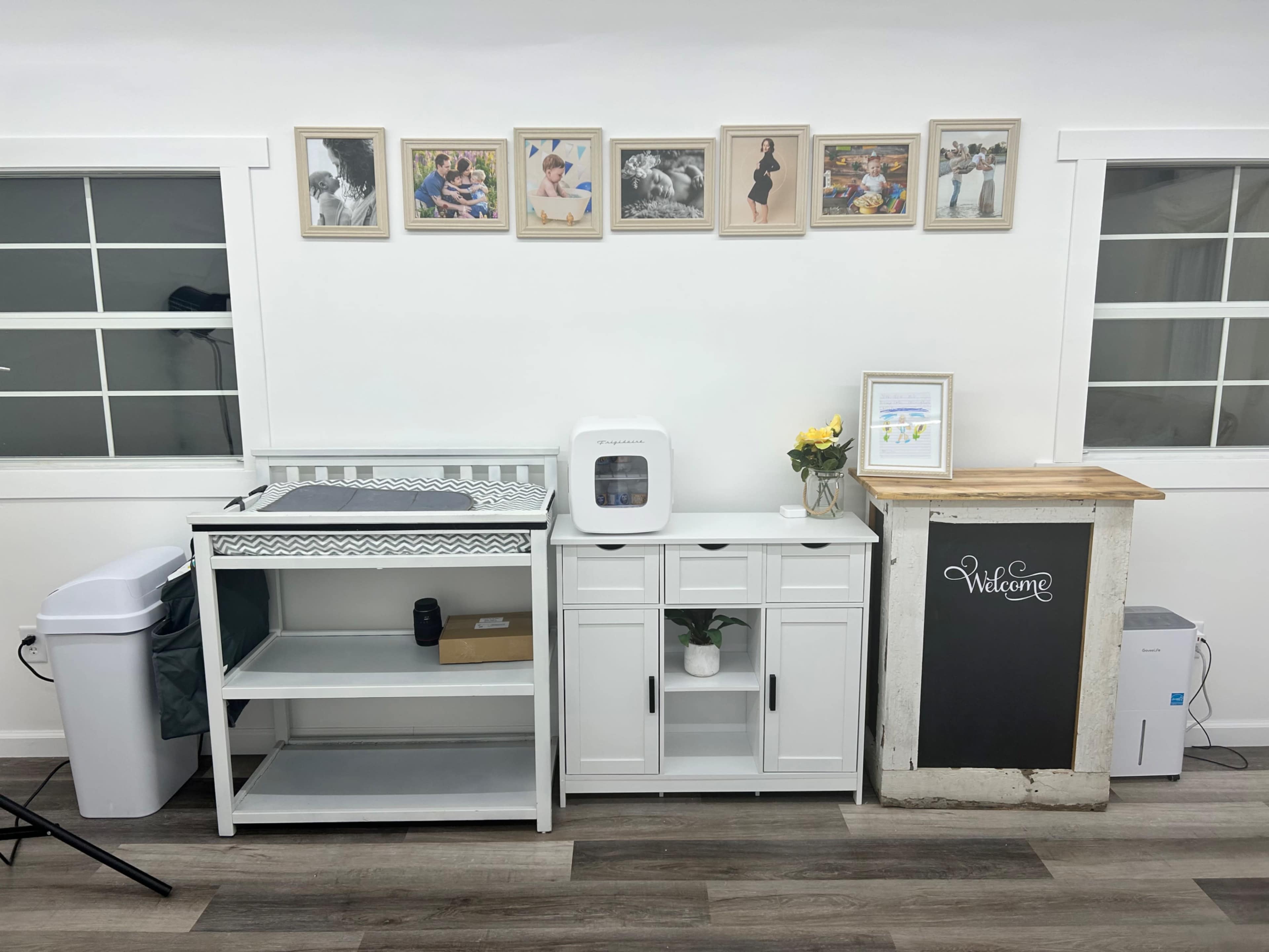 A clean white wall with a series of framed photos above a wooden reception desk, a small cabinet, and a changing table, all arranged in a well-organized space.
