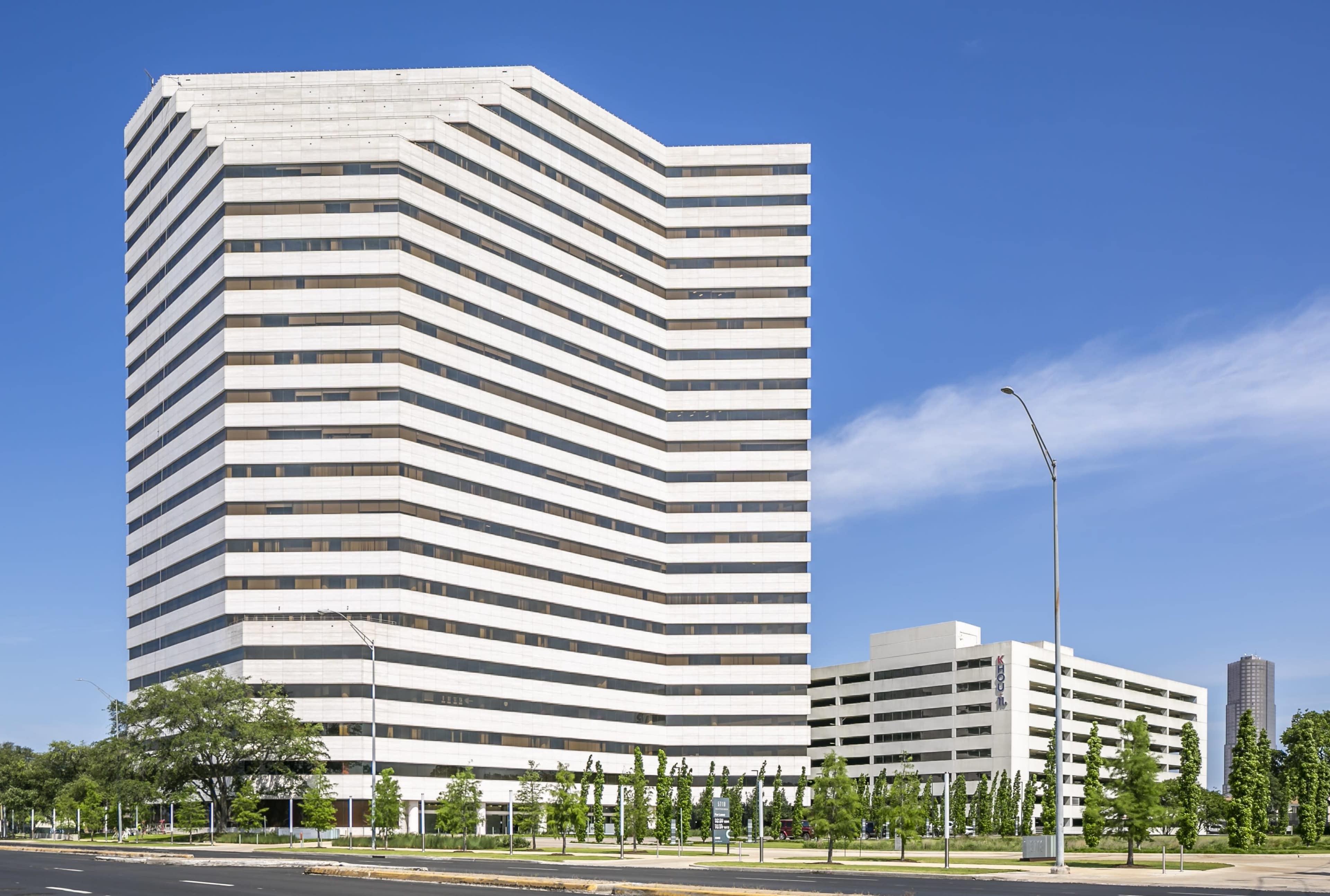 The image shows a modern multi-story office building with a striped façade and landscaped greenery in the foreground.