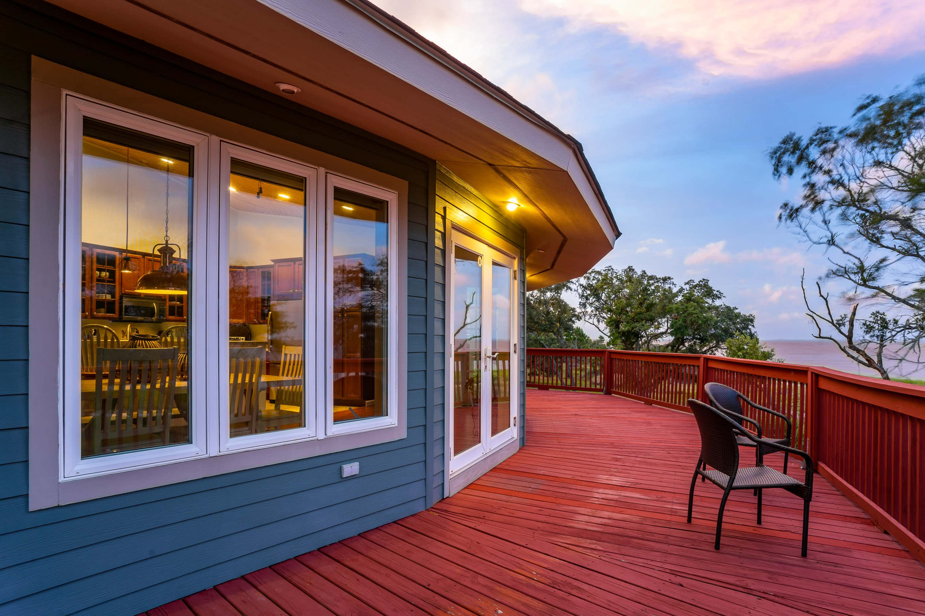 A wooden deck wraps around a blue house, featuring large windows and a single chair positioned near the railing.