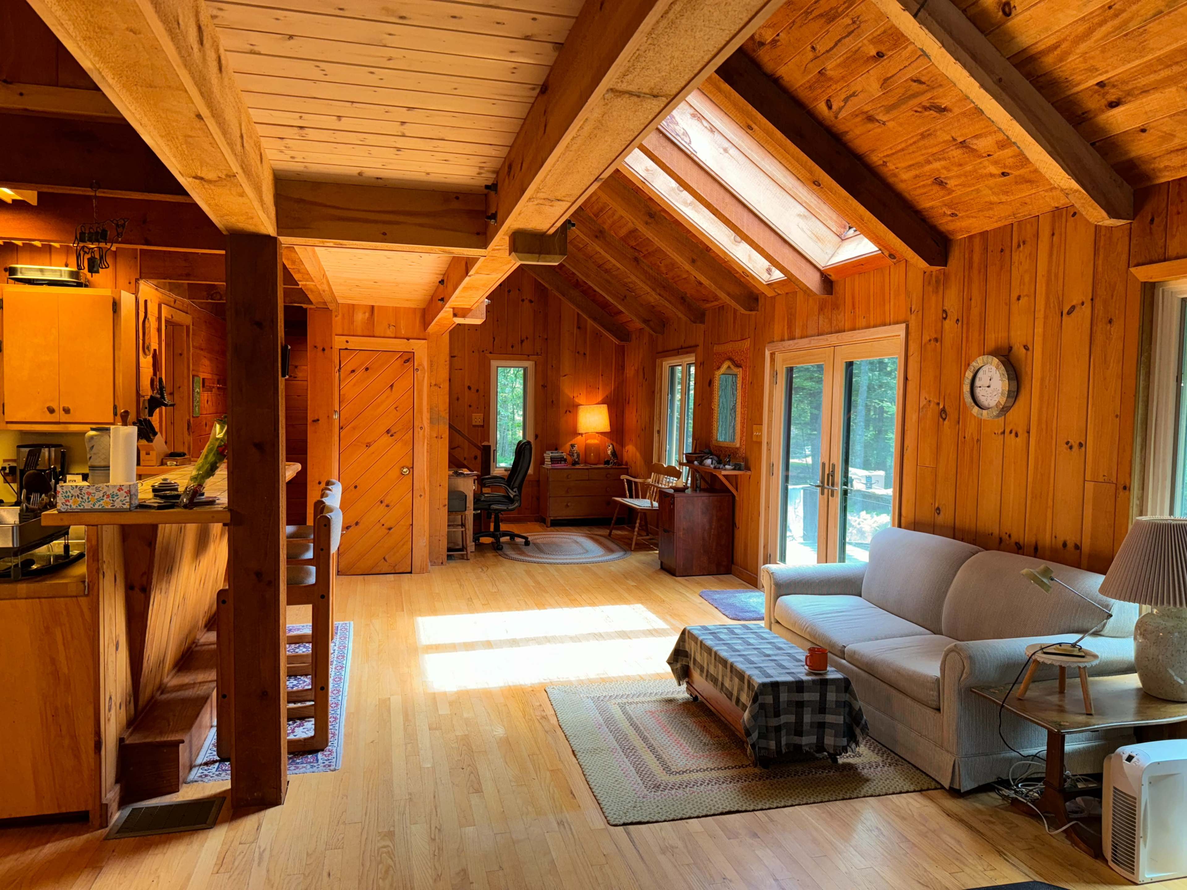 The image shows a spacious wooden interior of a cabin with a living area, kitchen, and workspace, featuring large windows that allow natural light to illuminate the space.