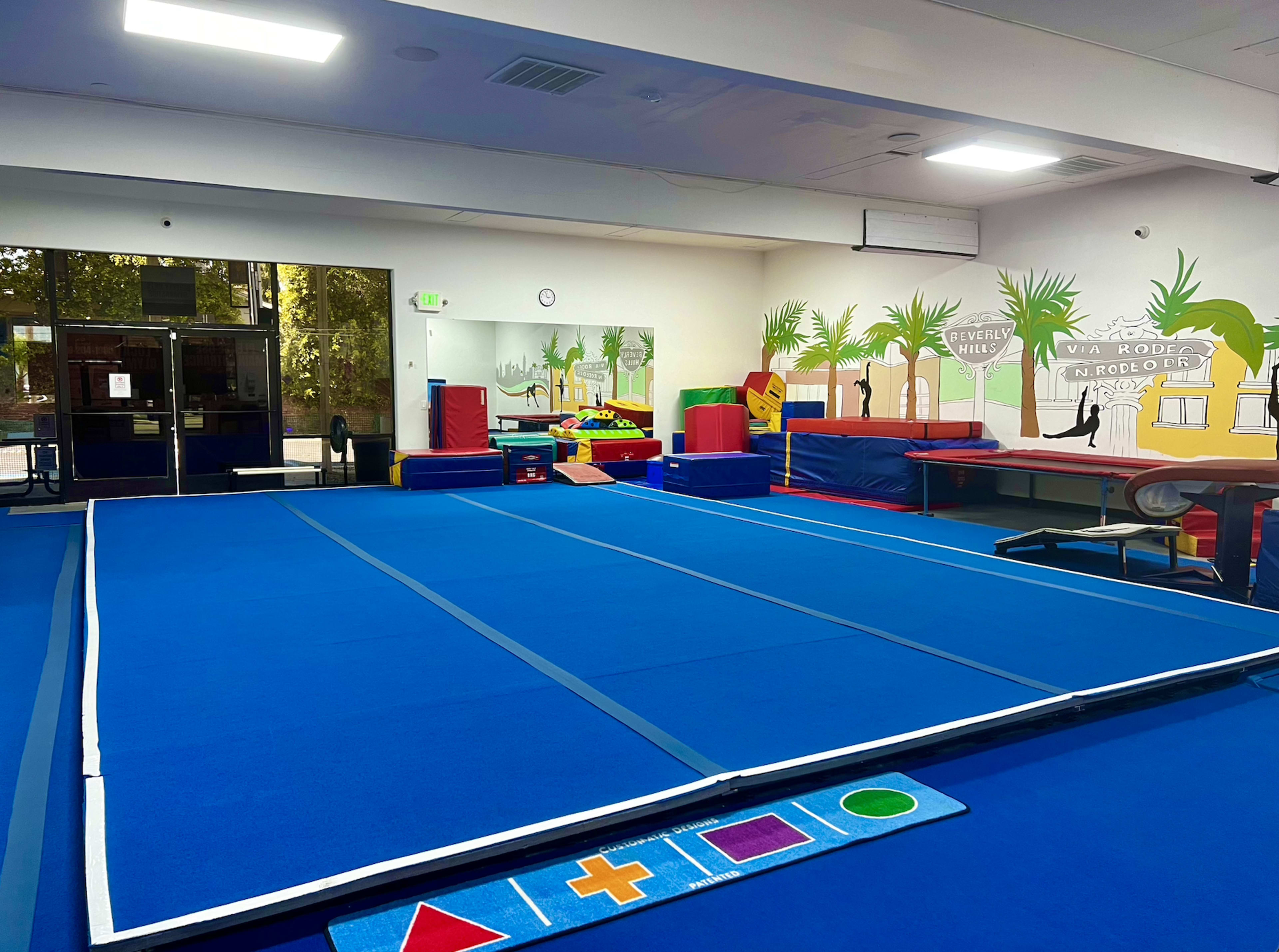 A gymnasium with a large blue tumbling mat, colorful exercise equipment, and a wall decorated with palm trees and playful shapes.