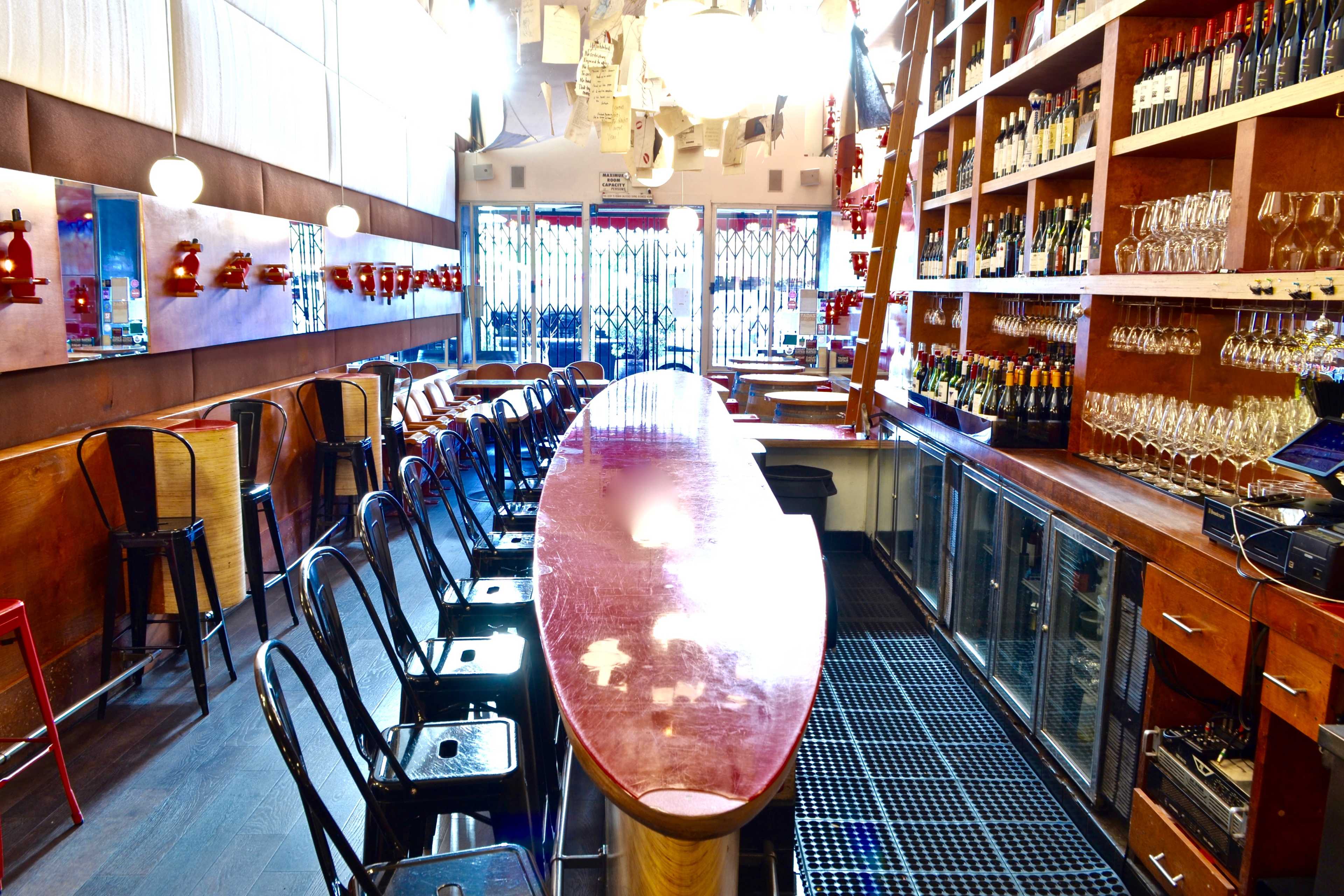 A bar area with an oval wooden counter, rows of empty chairs, and shelves stocked with various bottles and glasses.