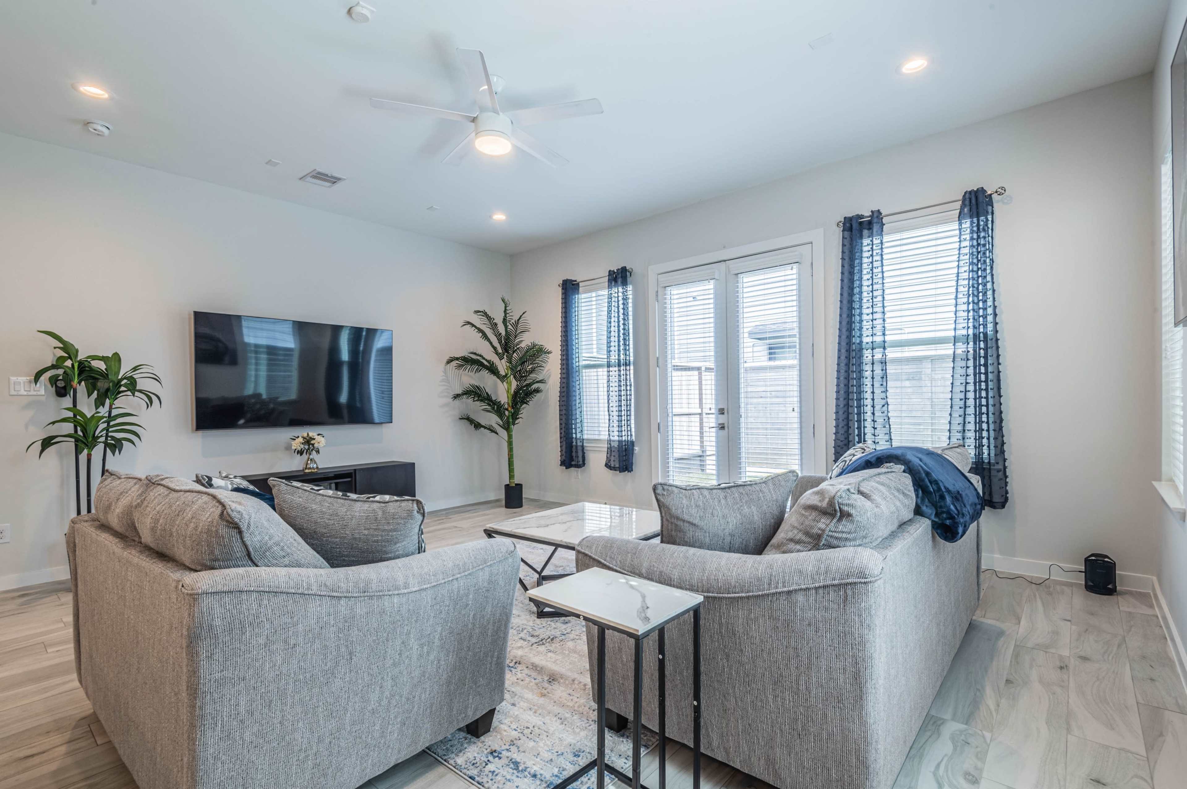 A modern living room features two gray sofas facing a coffee table, with a television mounted on the wall and large windows framed by blue curtains.