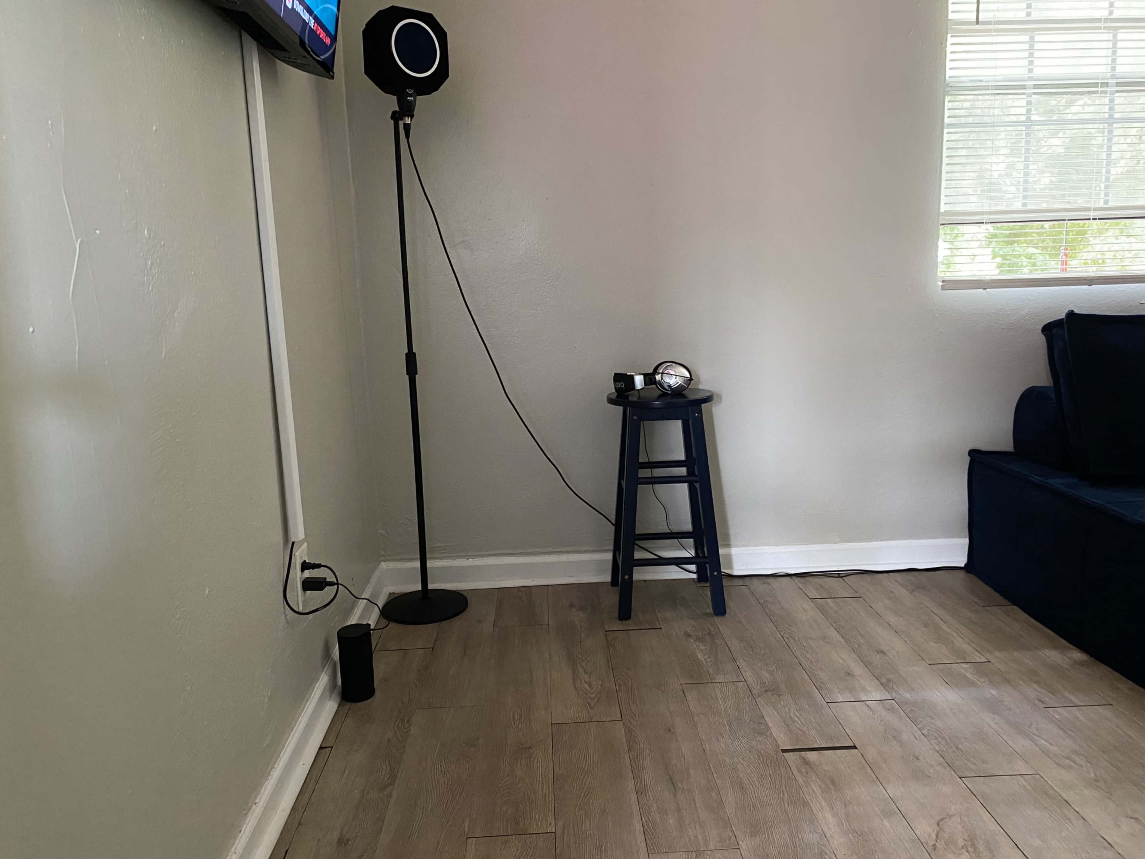 The image shows a corner of a room with a blue stool next to a black speaker, a standing lamp, and a dark blue sofa against a wall with a window.
