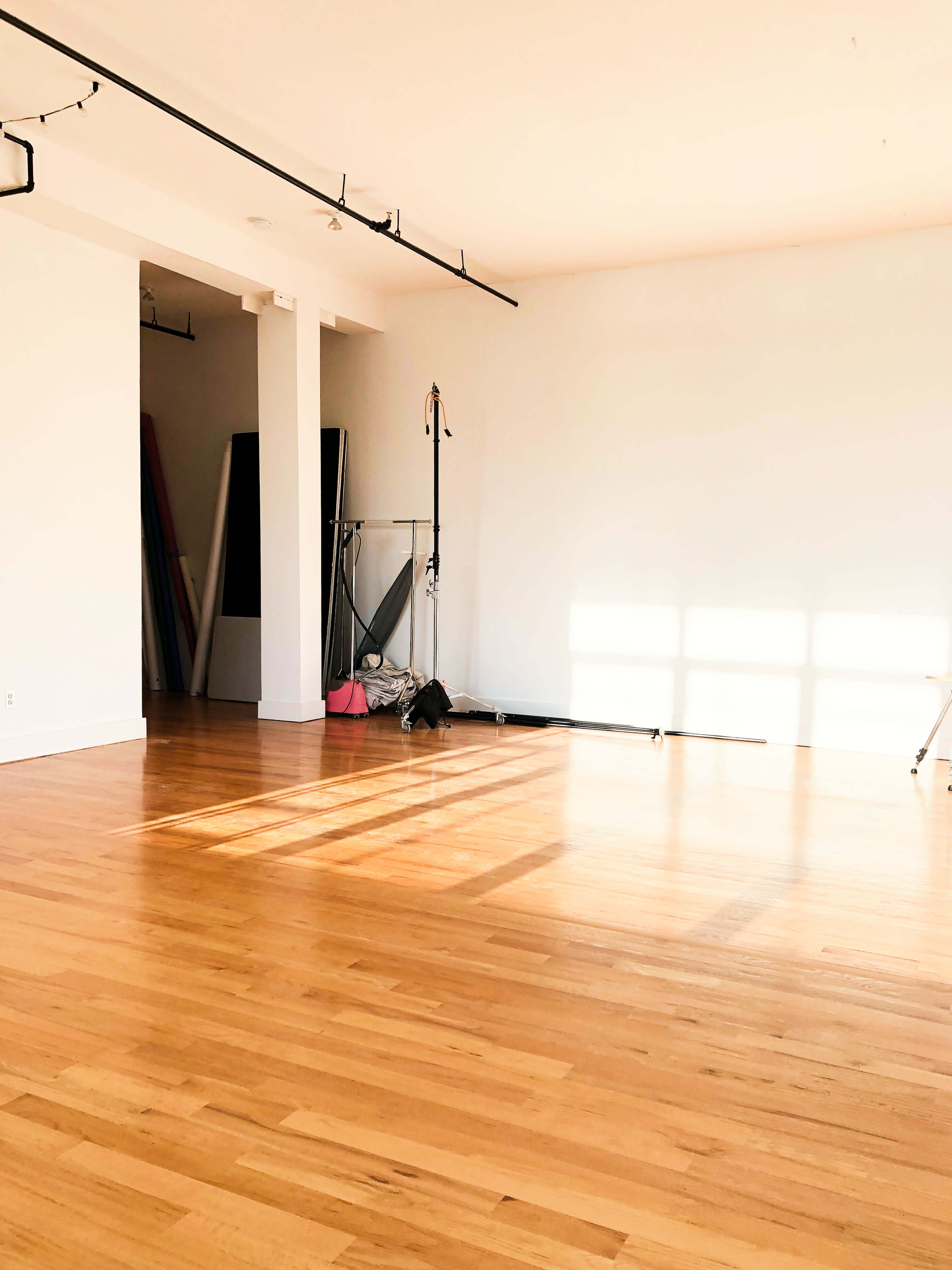 A spacious, empty room with wooden flooring and large windows allowing sunlight to cast shadows on the floor.