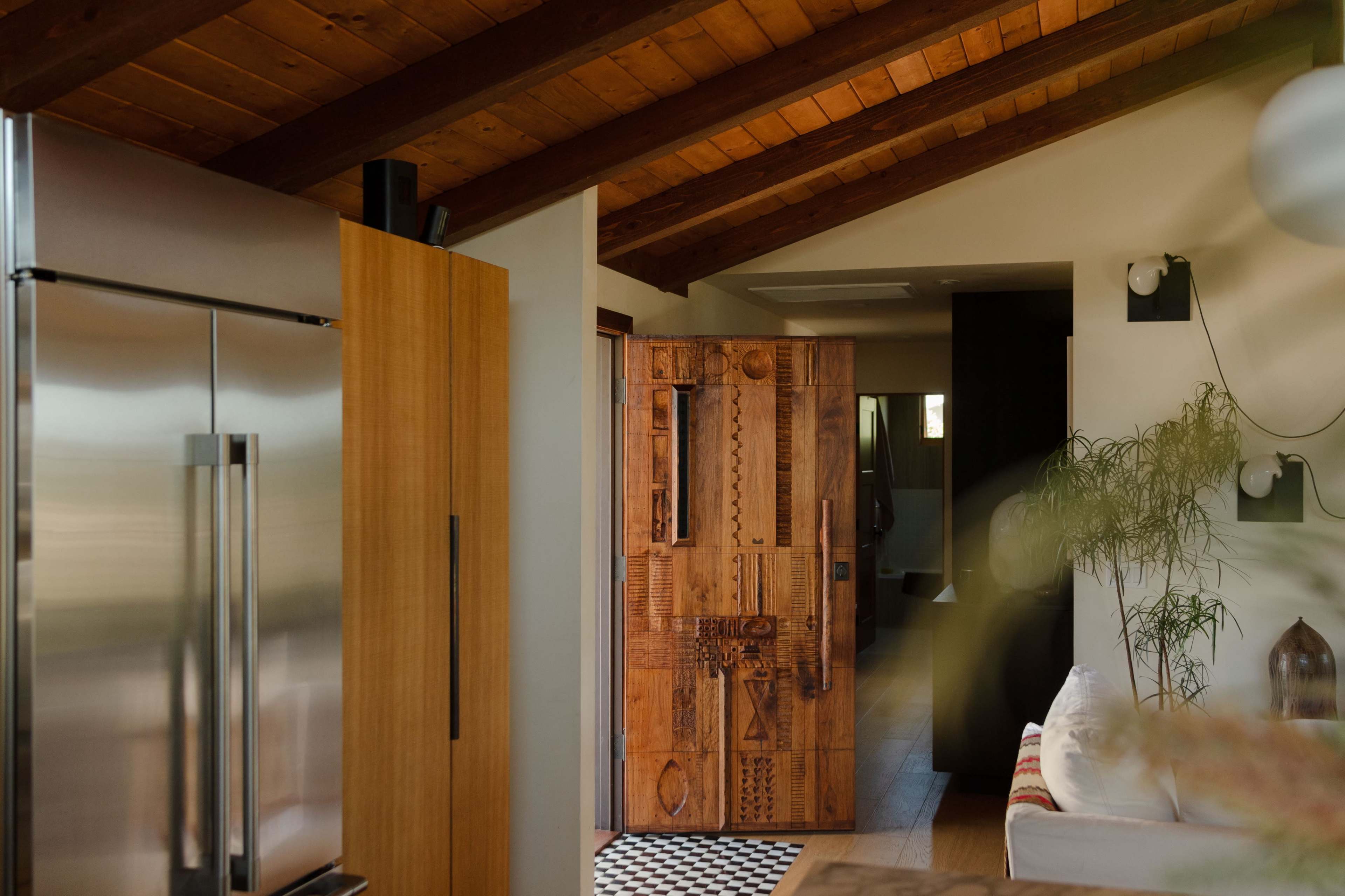 The image shows a modern kitchen featuring a stainless steel refrigerator, a wooden door with intricate carvings, and a view leading to a hallway with dark walls.