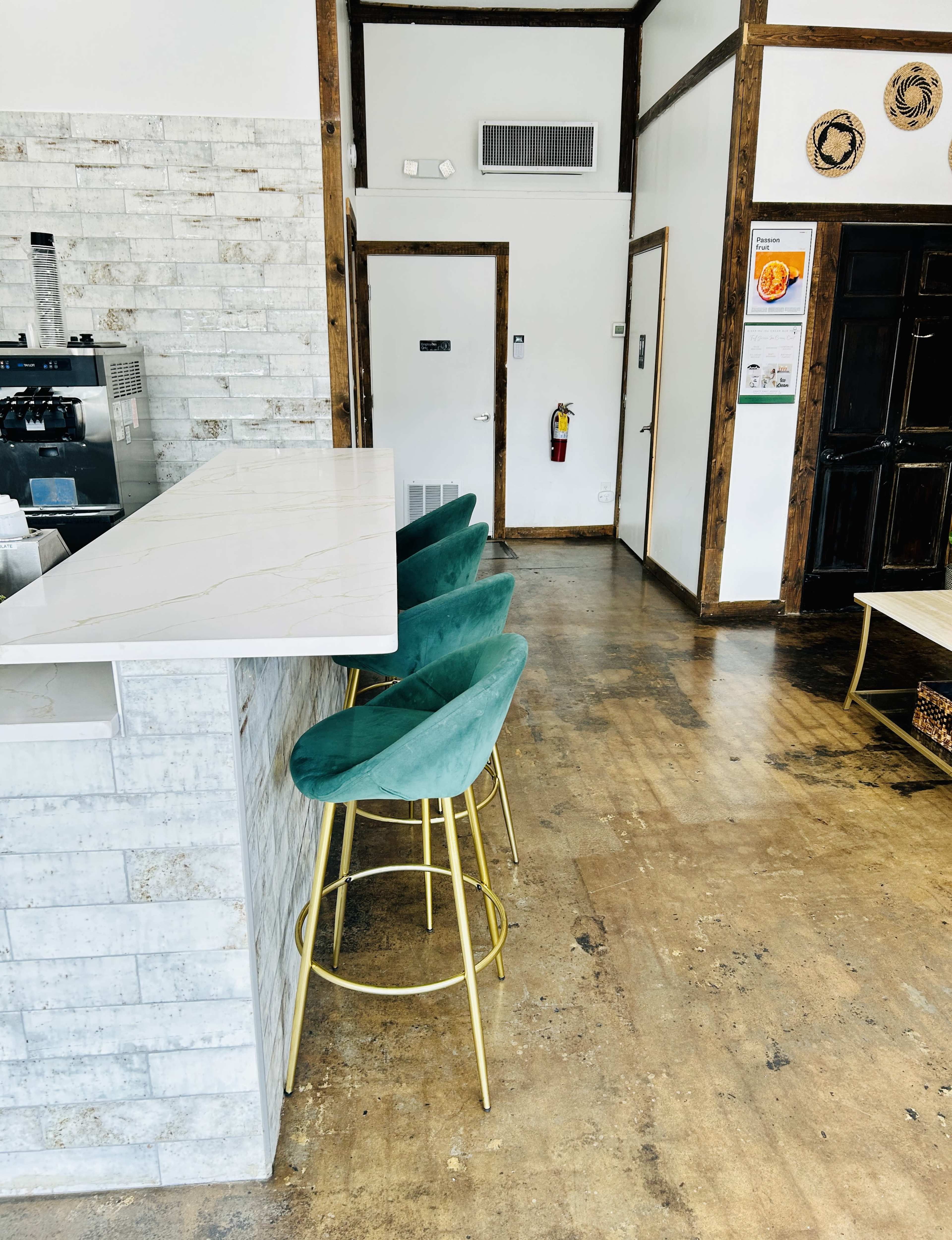 The image shows a coffee shop interior with a marble countertop, four green bar stools with gold legs, and a wooden floor.