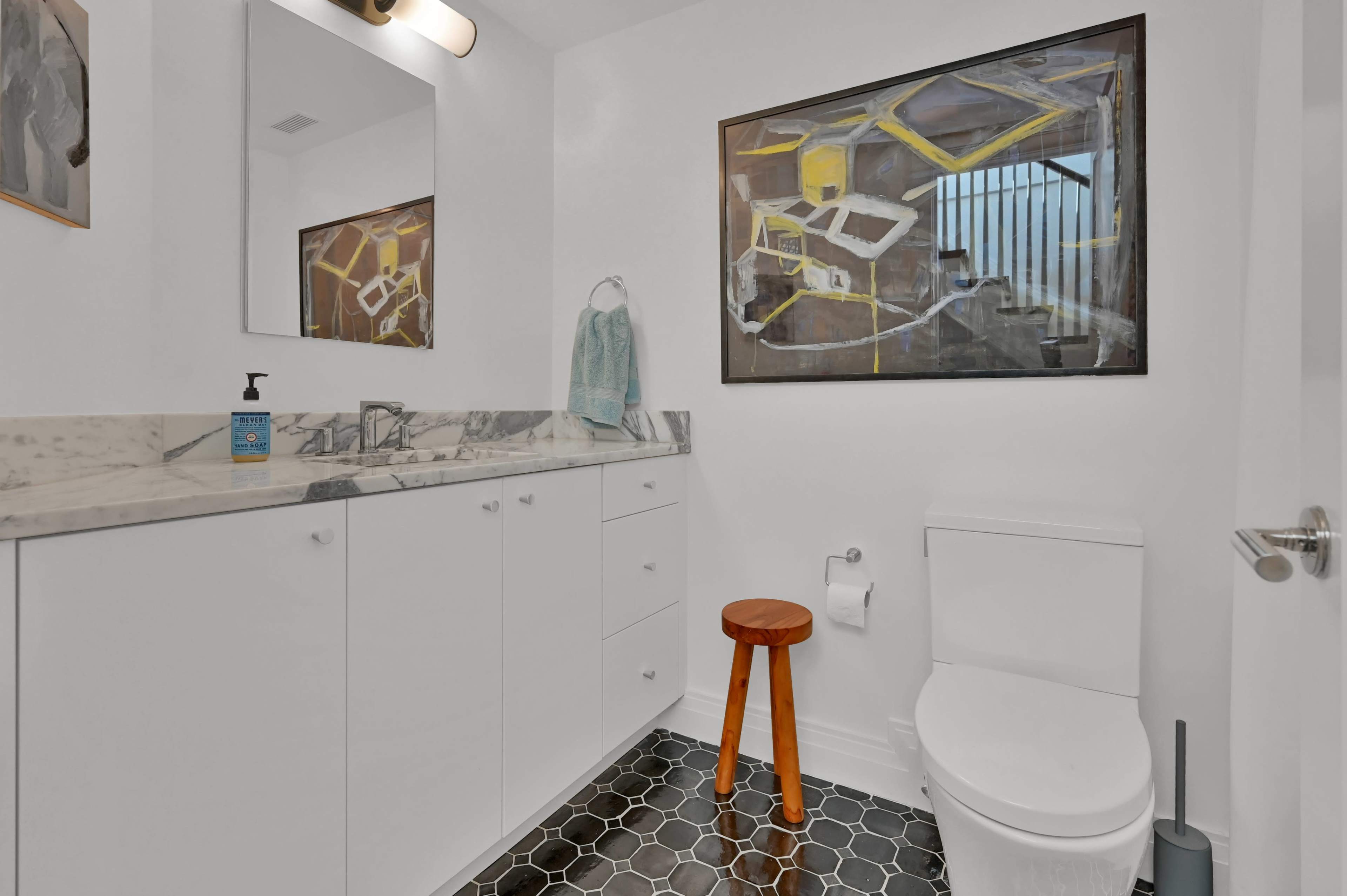 A modern bathroom featuring a white toilet, a marble countertop with a sink, a wooden stool, and a framed abstract painting on the wall.