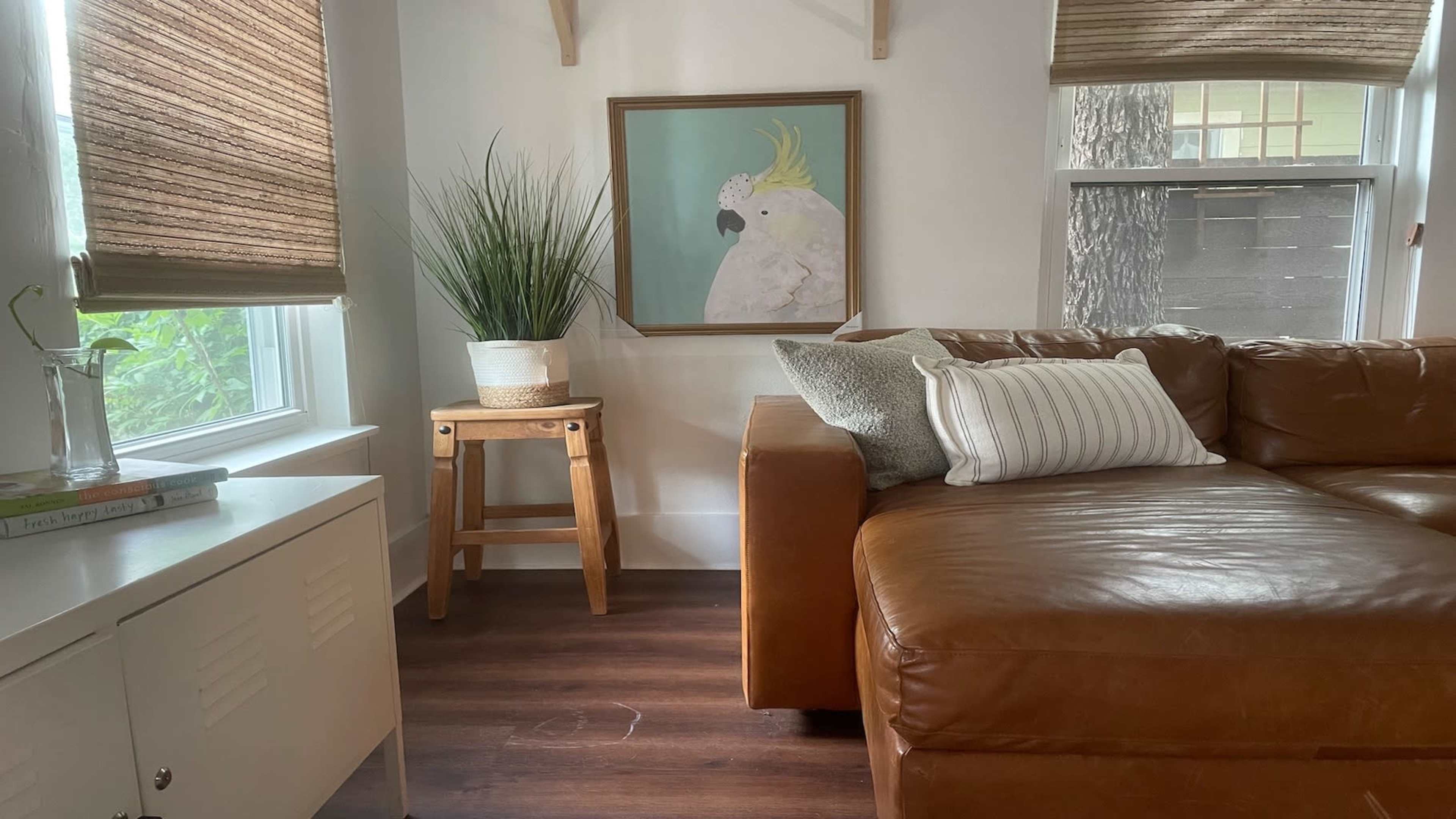A cozy living room corner featuring a brown leather sofa, a small wooden stool with a potted plant, and a framed artwork of a bird on the wall.