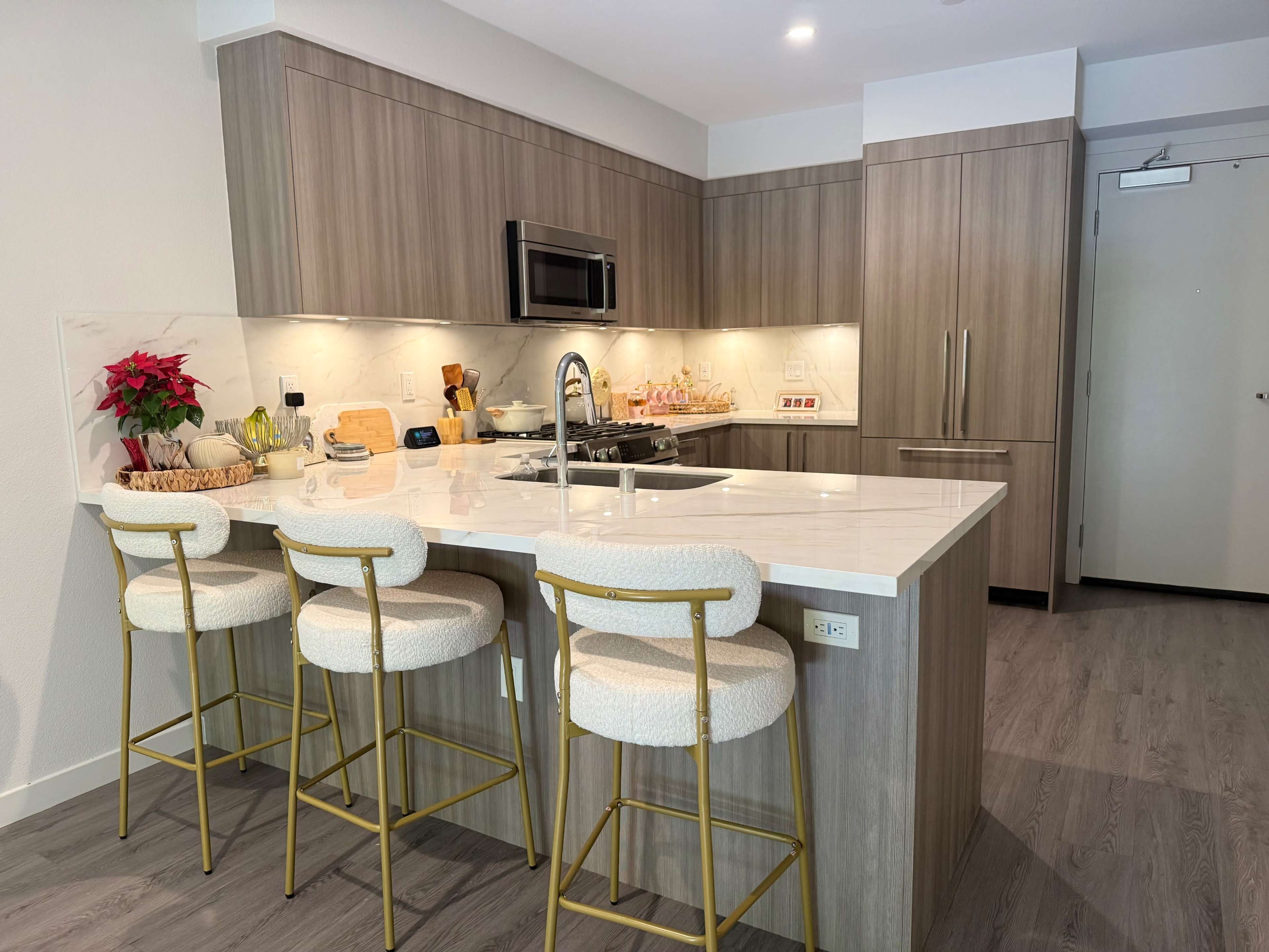 The image shows a modern kitchen featuring wooden cabinetry, a central island with bar stools, and various appliances.