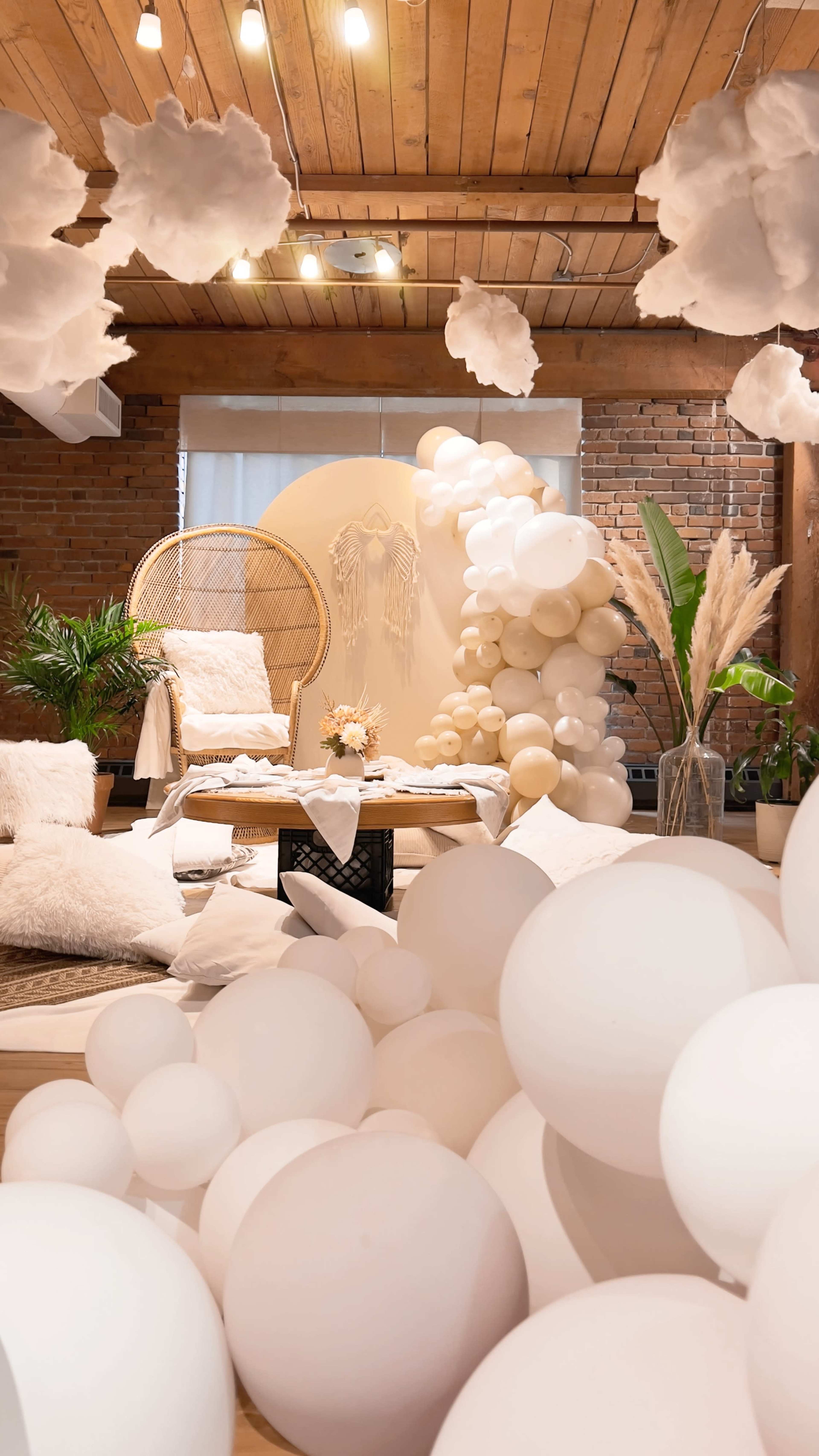 A decorated interior space features a cozy seating area with a rattan chair, a backdrop of balloons, and white cloud-like decorations hanging from the ceiling.