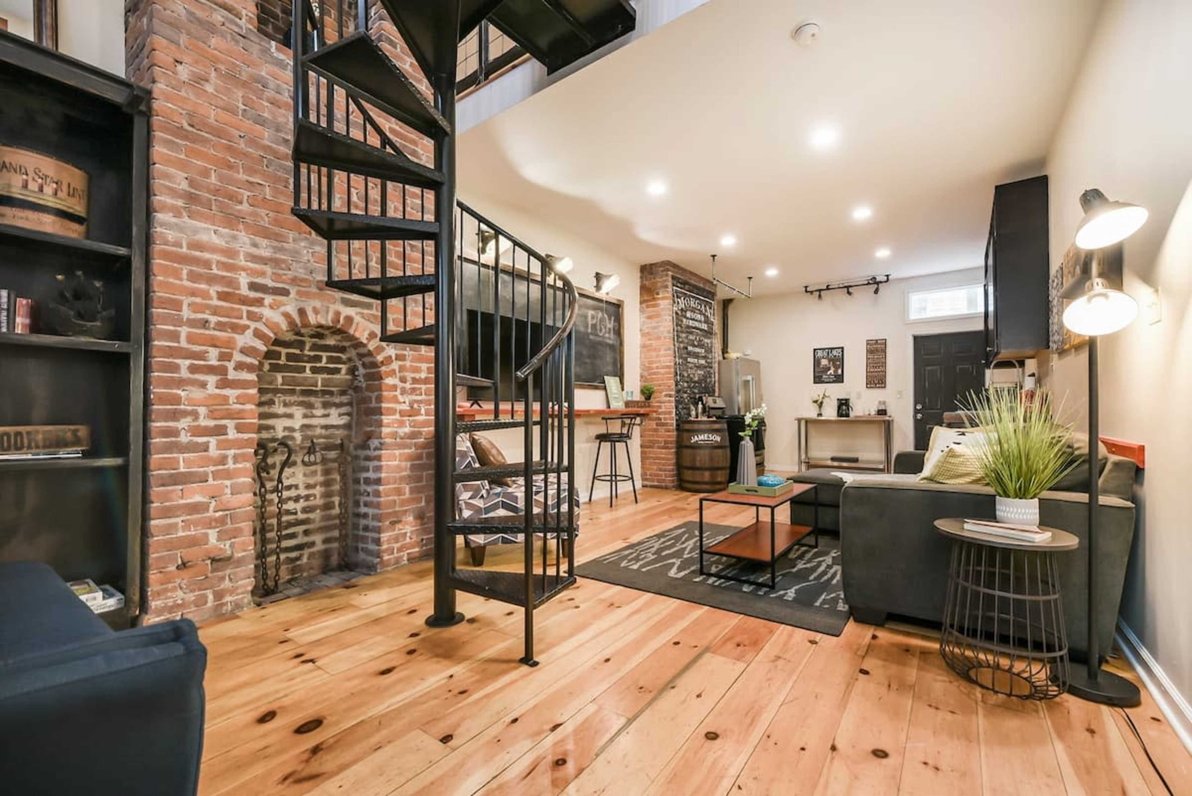 A cozy living space featuring exposed brick walls, a spiral staircase, and wooden flooring, with a seating area and a small bar.